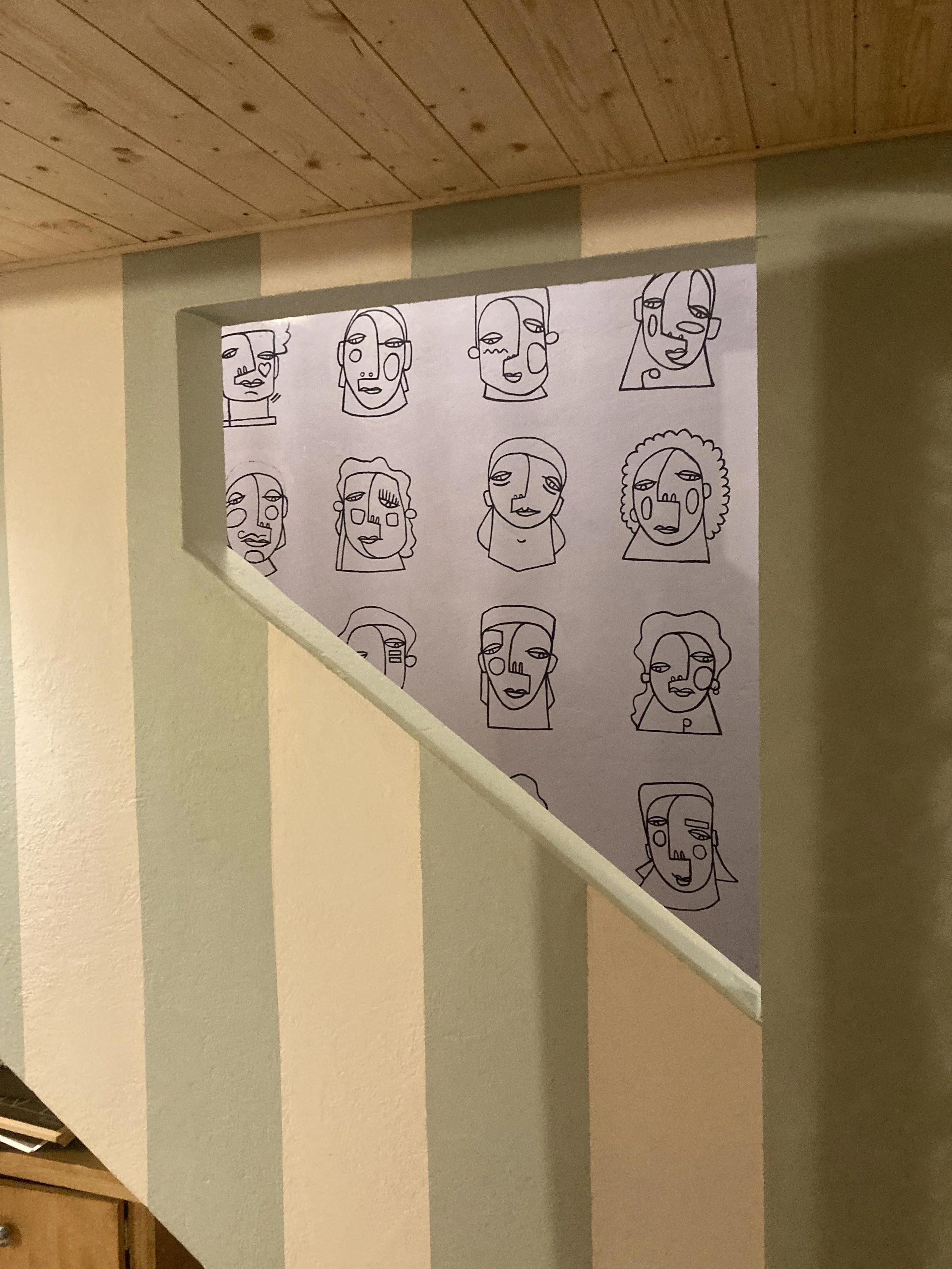 Dessin de plusieurs visages de style abstrait ou caricatural, réalisés avec des lignes continues, affichés sur un panneau fixé dans un intérieur avec un mur peint à rayures et un plafond en bois.