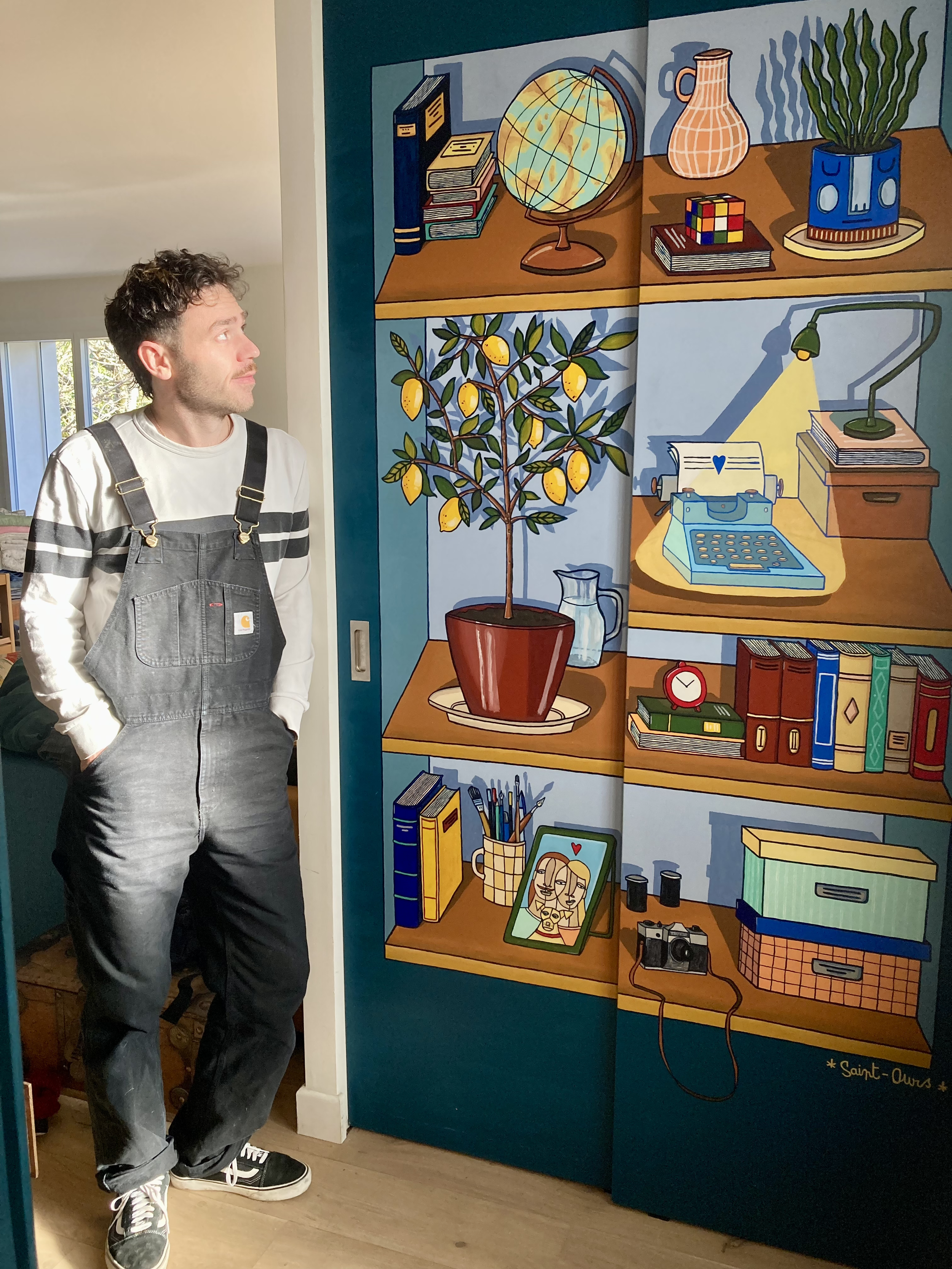 Un homme se tient devant un mur peint avec une étagère illustrée contenant une plante en pot, une globe terrestre, un Rubik's Cube, une machine à écrire, un livre, une caméra, des boîtes de rangement et une photo encadrée.