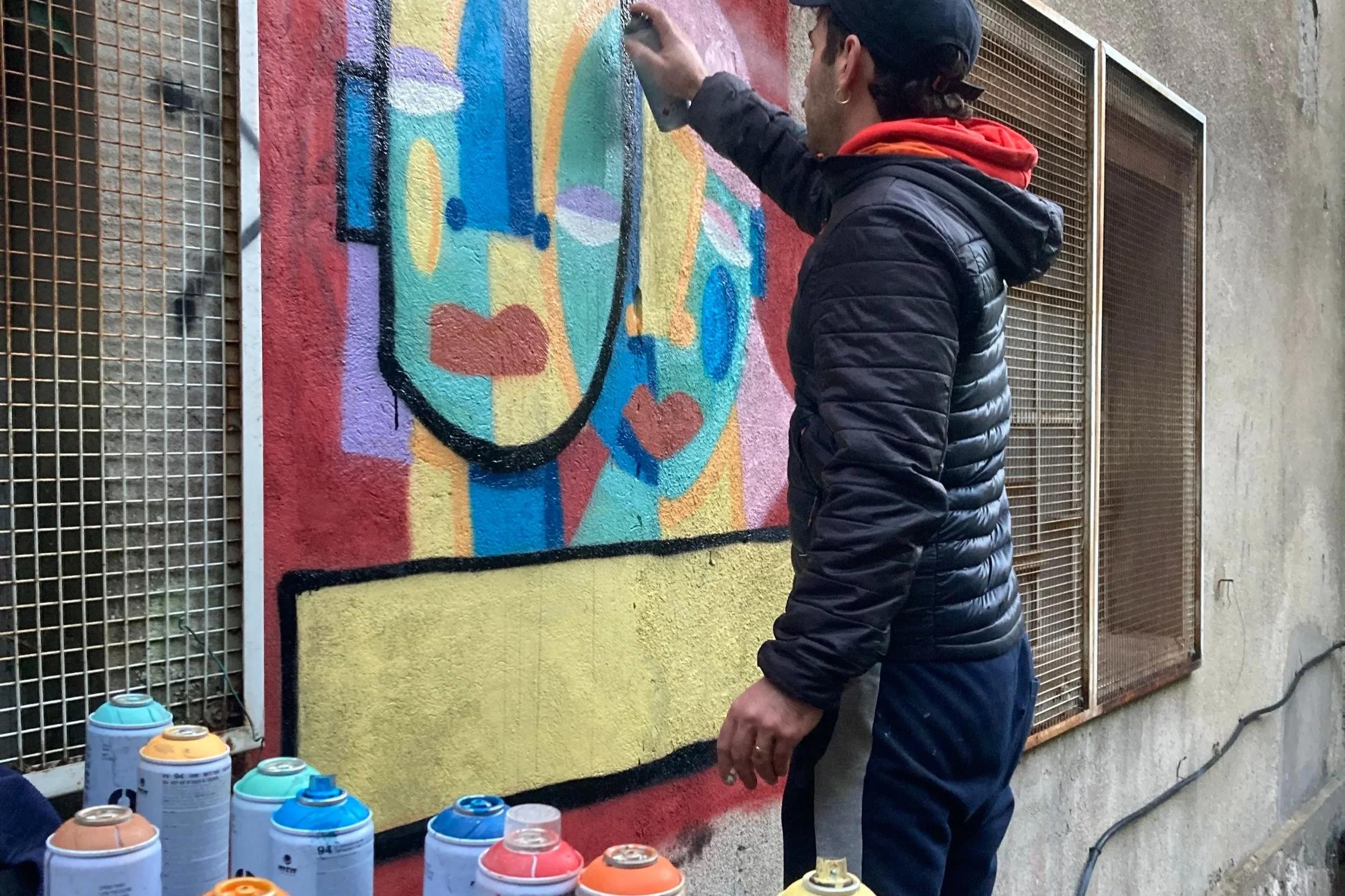 Un homme en vest jacket noir, casquette, et sweat à capuche rouge, en train de faire un graffiti coloré sur un mur extérieur.