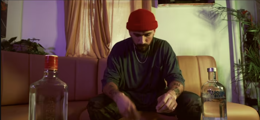 Young man with a beard, red beanie, black shirt, and tattoos, sitting on a beige sofa with two bottles of alcohol in front of him, in a dimly lit room with plants and curtains.
