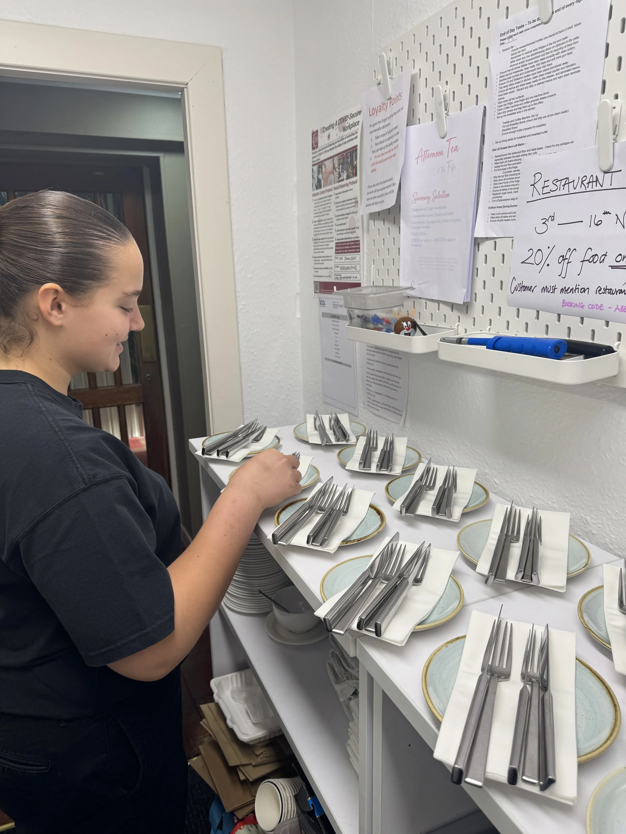 A woman standing by a table with neatly arranged silverware on plates and white napkins, preparing for a meal in a room with informational posters and papers on a white pegboard wall.