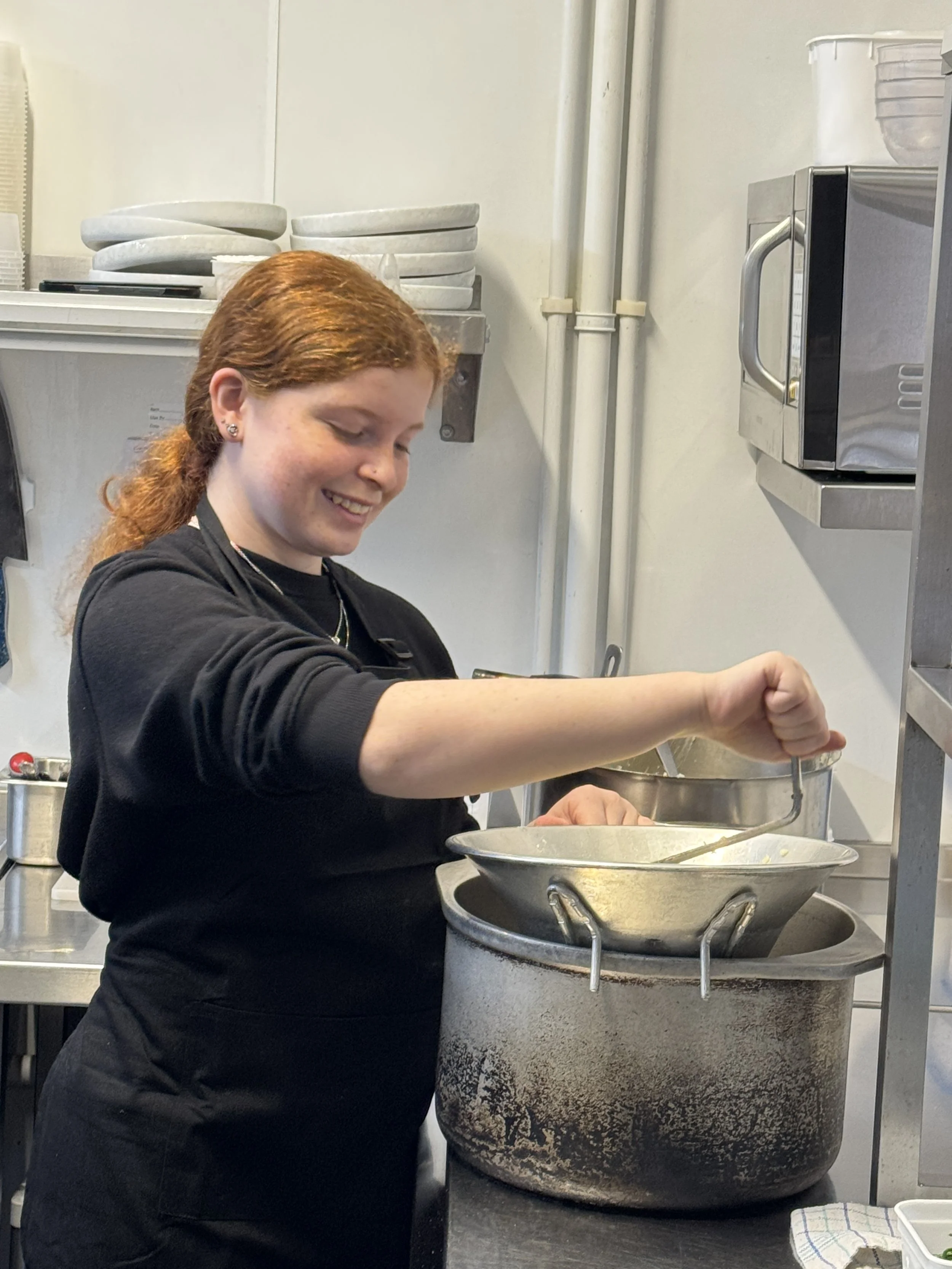 A young woman with red hair and a black shirt is cooking or working in a commercial kitchen, stirring or preparing food in a large pot on a stove.