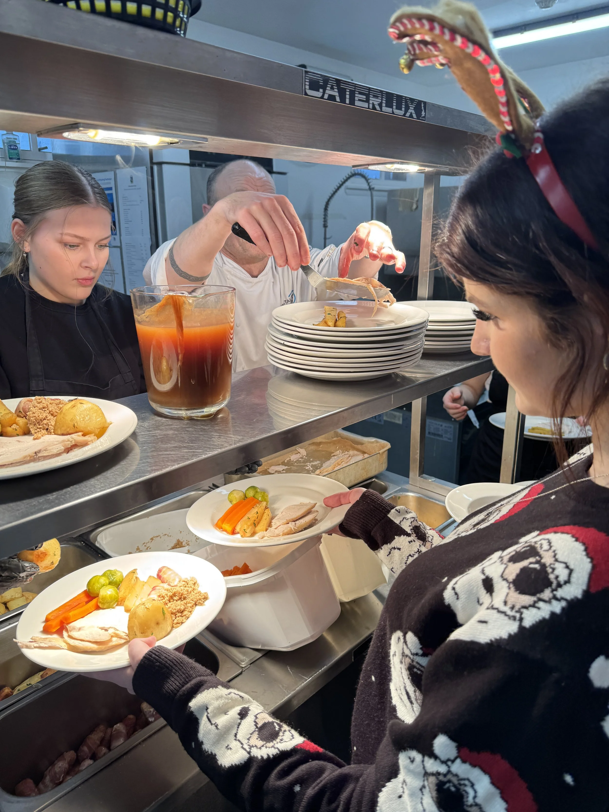 A person in a sweater with snowflake and reindeer patterns is serving food to customers at a cafeteria-style line, with plates of cooked vegetables and meat visible.