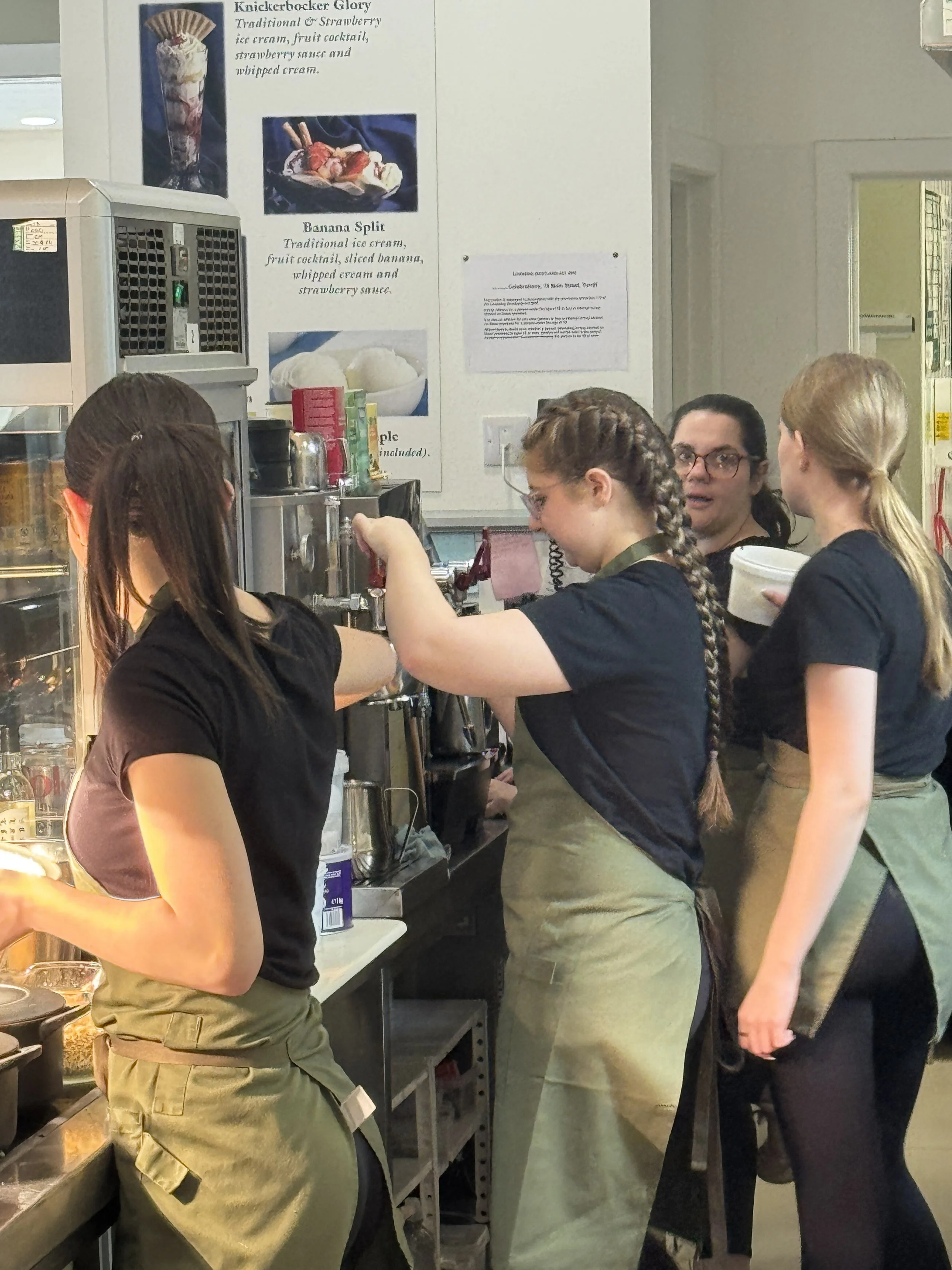 A young woman in a white shirt and black apron is garnishing a salad with a squeeze bottle in a professional kitchen.