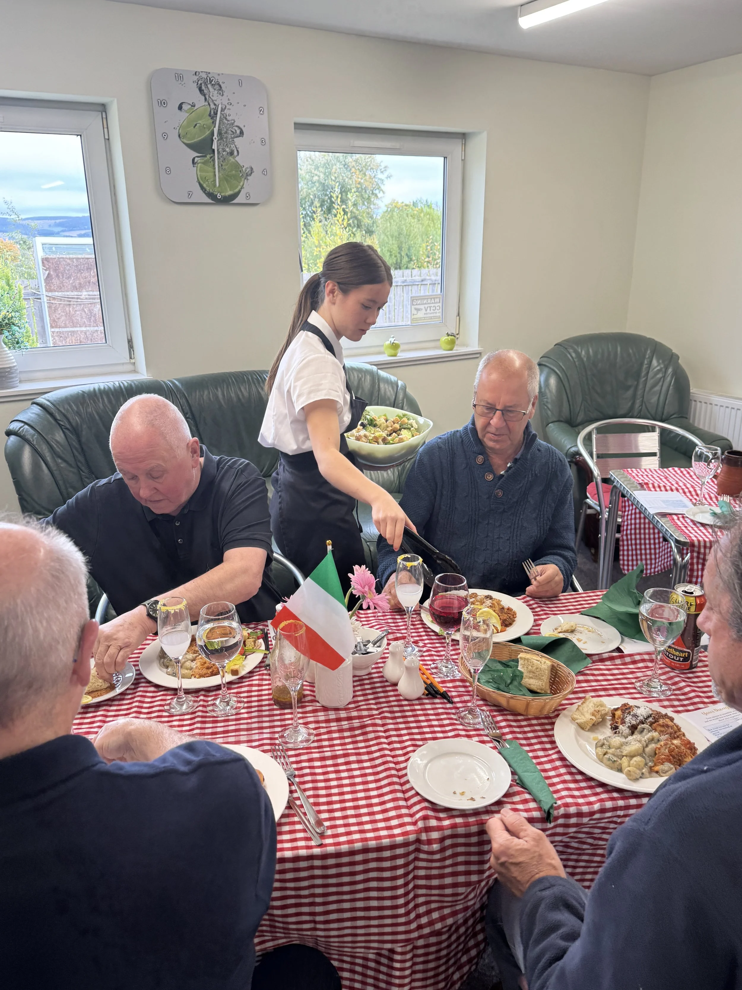 A group of people sitting around a table with red and white checkered tablecloth, enjoying a meal. A young woman is serving food to a man, and there are plates of food, drinks, and condiments on the table. The setting appears to be a cozy dining room with windows showing a view of the outdoors, and a wall clock above the window.