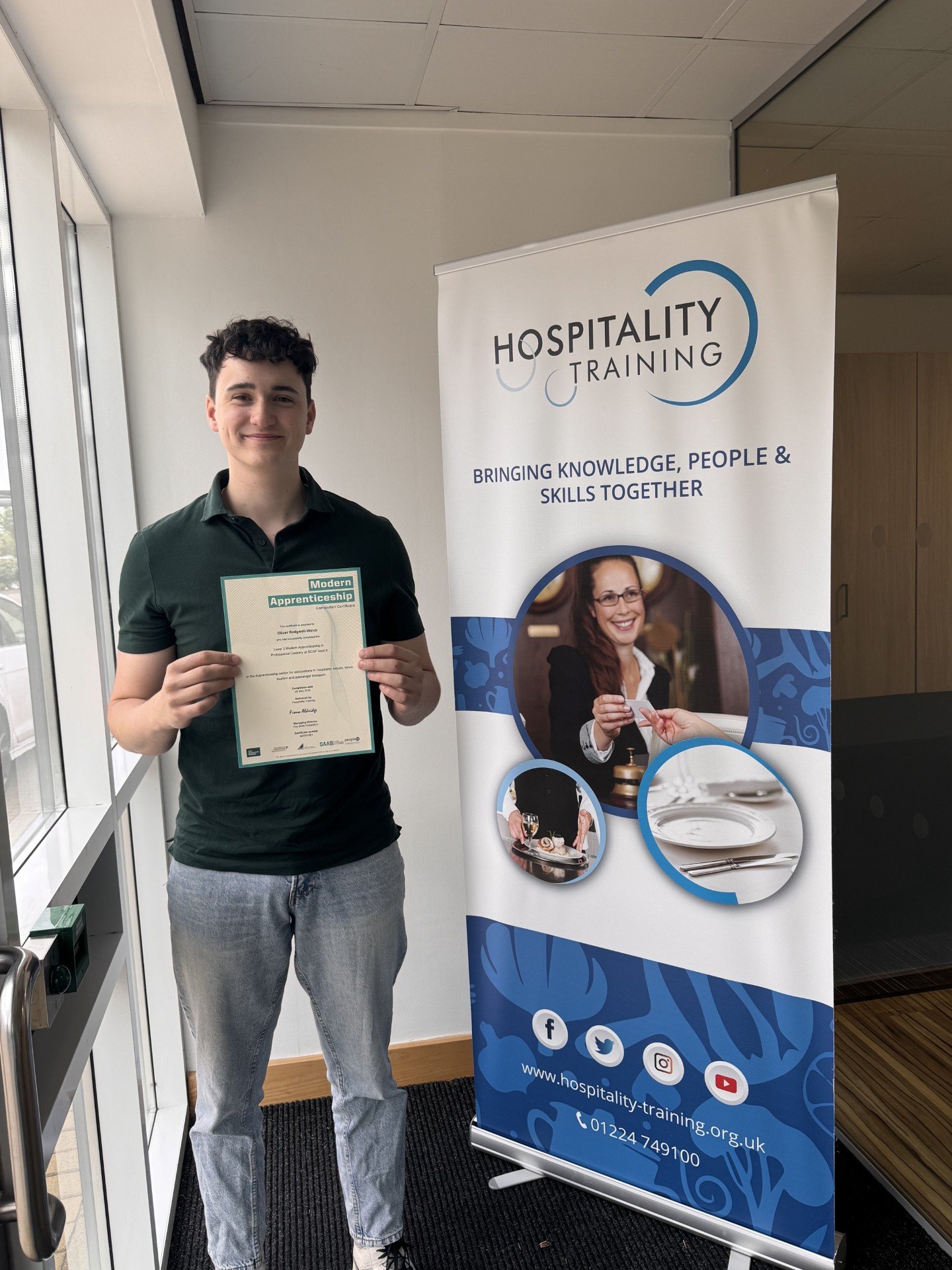 A young man in a chef uniform and striped apron holding a certificate for a Modern Apprenticeship standing in front of a poster with people working in a kitchen. Modern Apprenticeships Hospitality Training Aberdeen Scotland