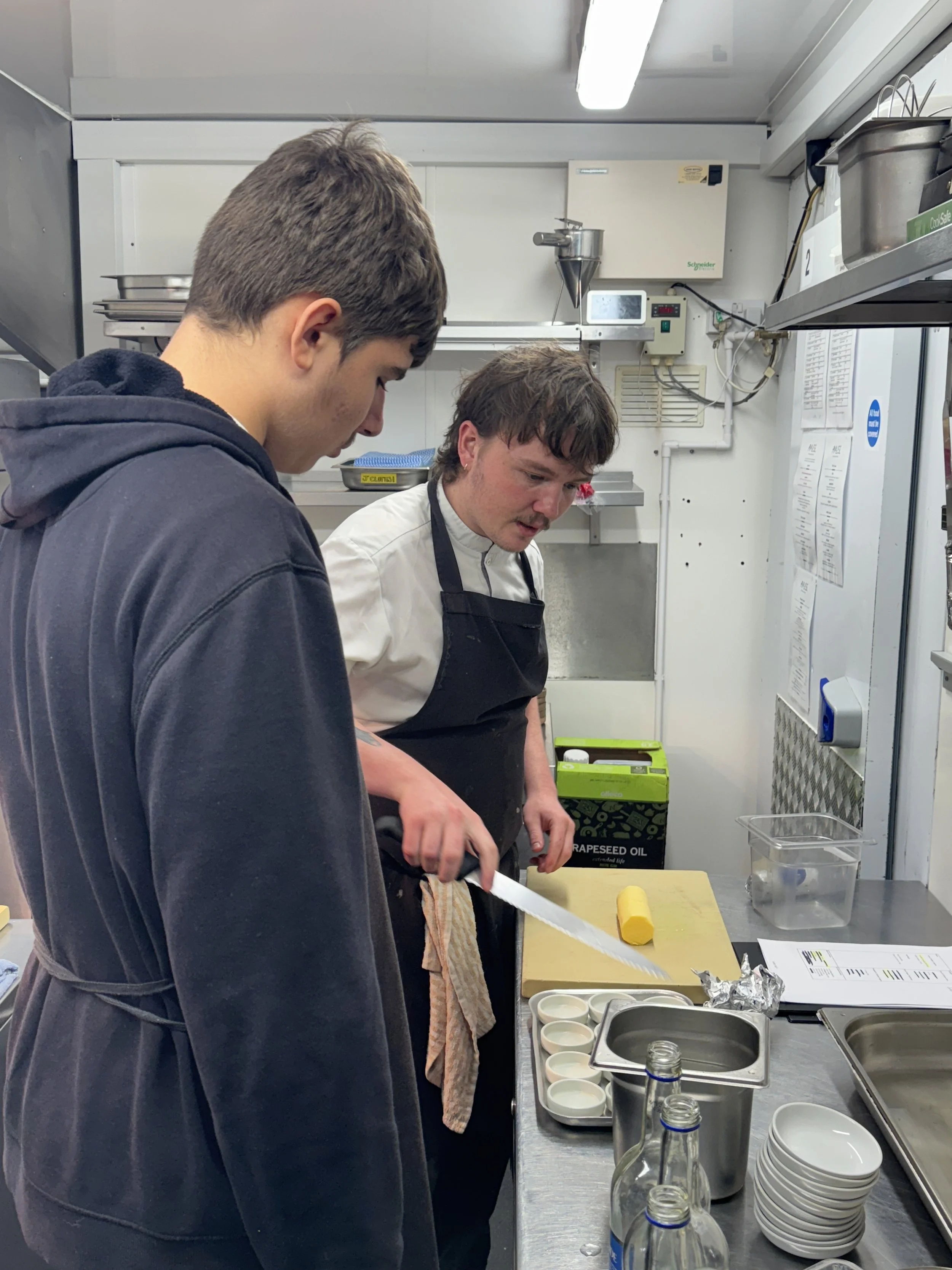 Two men in a kitchen preparing food; one man is slicing a yellow block of butter or similar ingredient on a cutting board, while the other is standing nearby observing. The kitchen has shelves with bowls, jars, and kitchen equipment, and a stainless steel countertop with various kitchen utensils and containers.