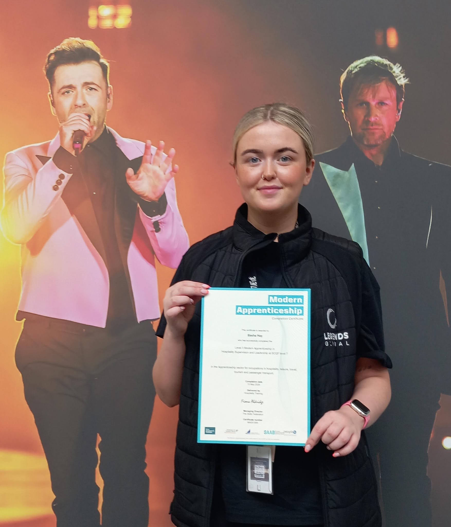 A young woman with blonde hair holding a certificate for a modern apprenticeship at an event, standing in front of a colorful poster of a singer with a microphone.