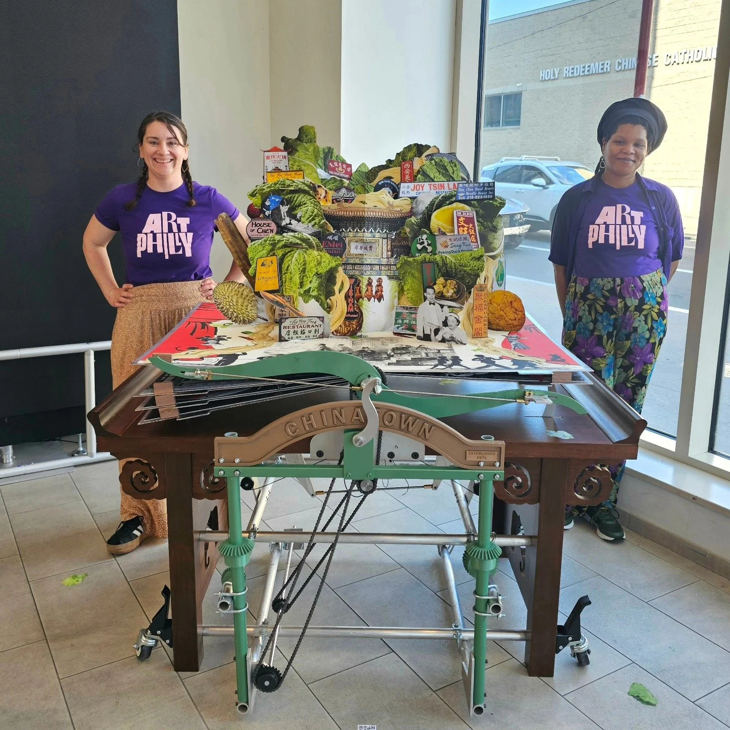 Two women standing beside a table with a colorful arrangement of various items and decorations inside a room with a window showing a parking lot outside.