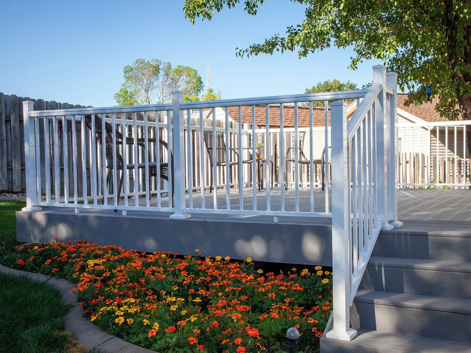 Backyard deck with white railings, gray flooring, and black chairs, surrounded by colorful flowers and grass, with a wooden fence and house in the background.