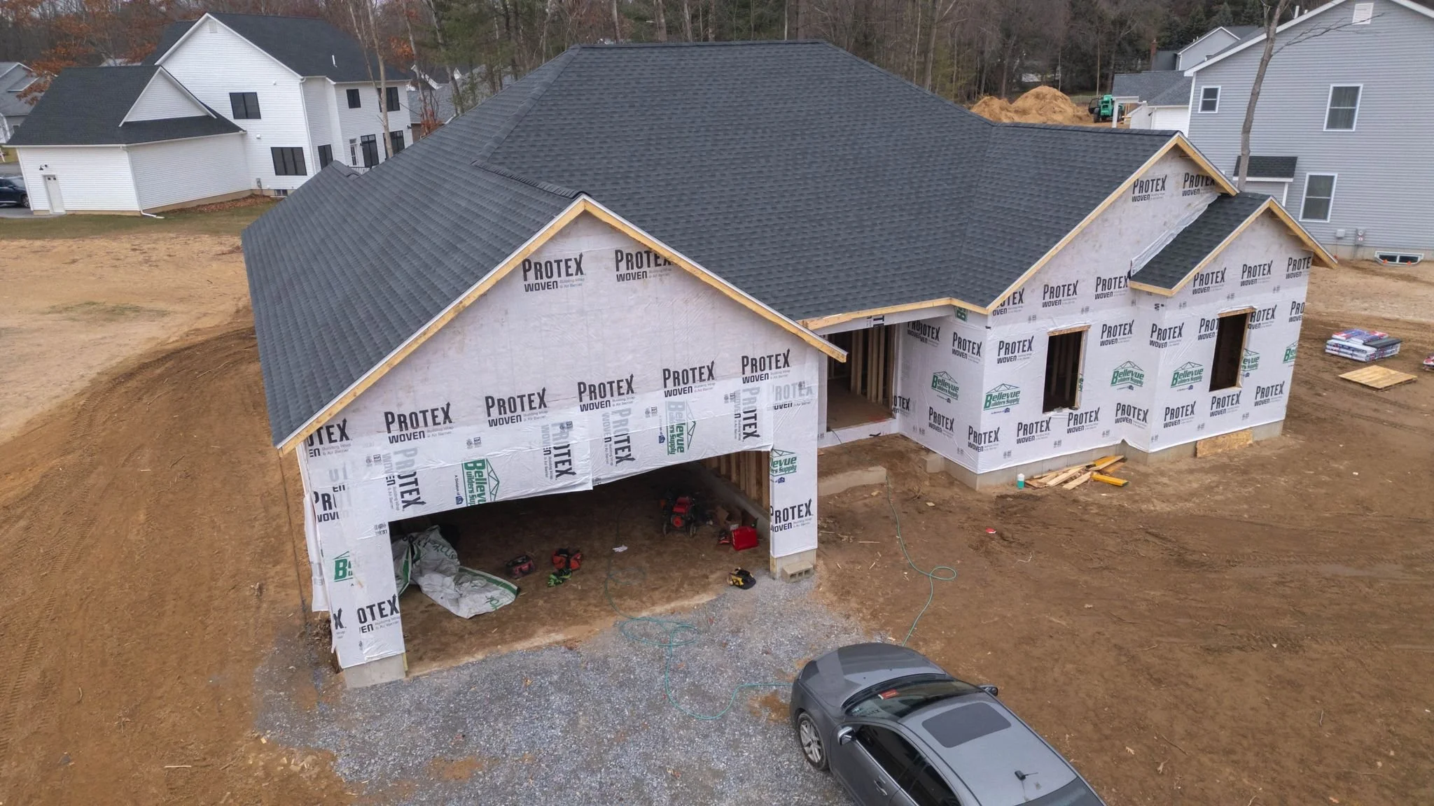A house under construction with the exterior wrapped in Protective house wrap and a completed shingled roof. The house is built into a slight hill with open garage space, and a silver car is parked in the gravel driveway. Surrounding houses are visible in the background.