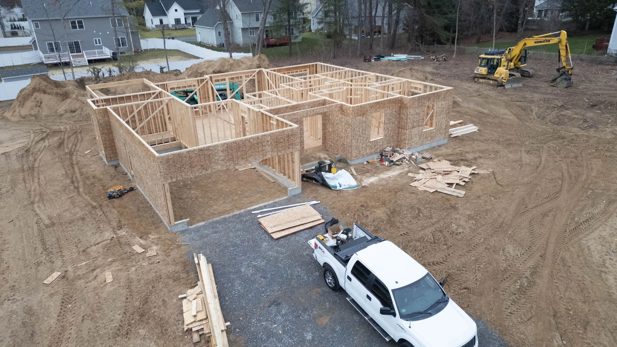 A house under construction with wooden framing, situated on a dirt lot with construction equipment and materials around.