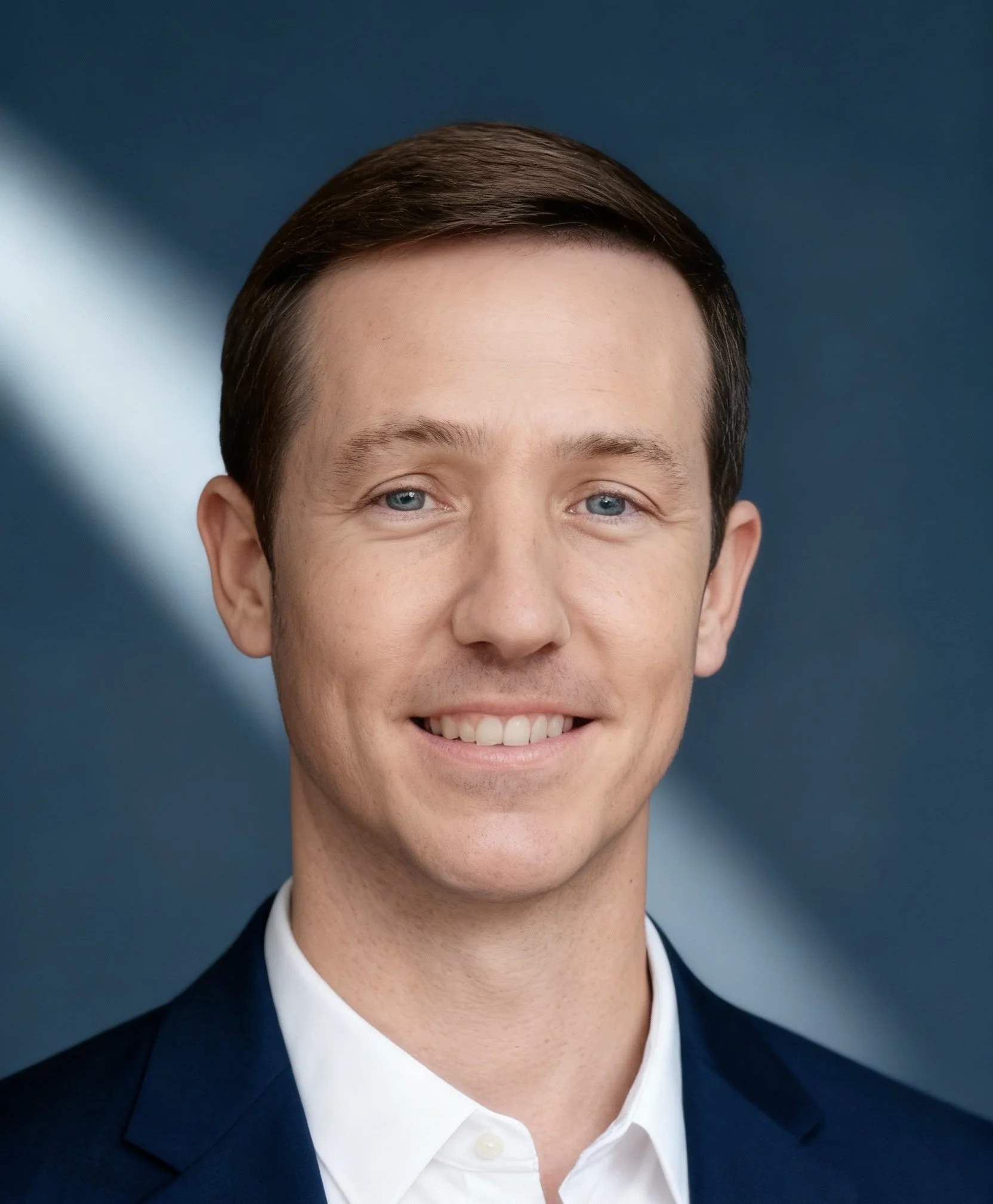 Man smiling outdoors, wearing a blue jacket and patterned shirt, with a wooden background and some branches visible.