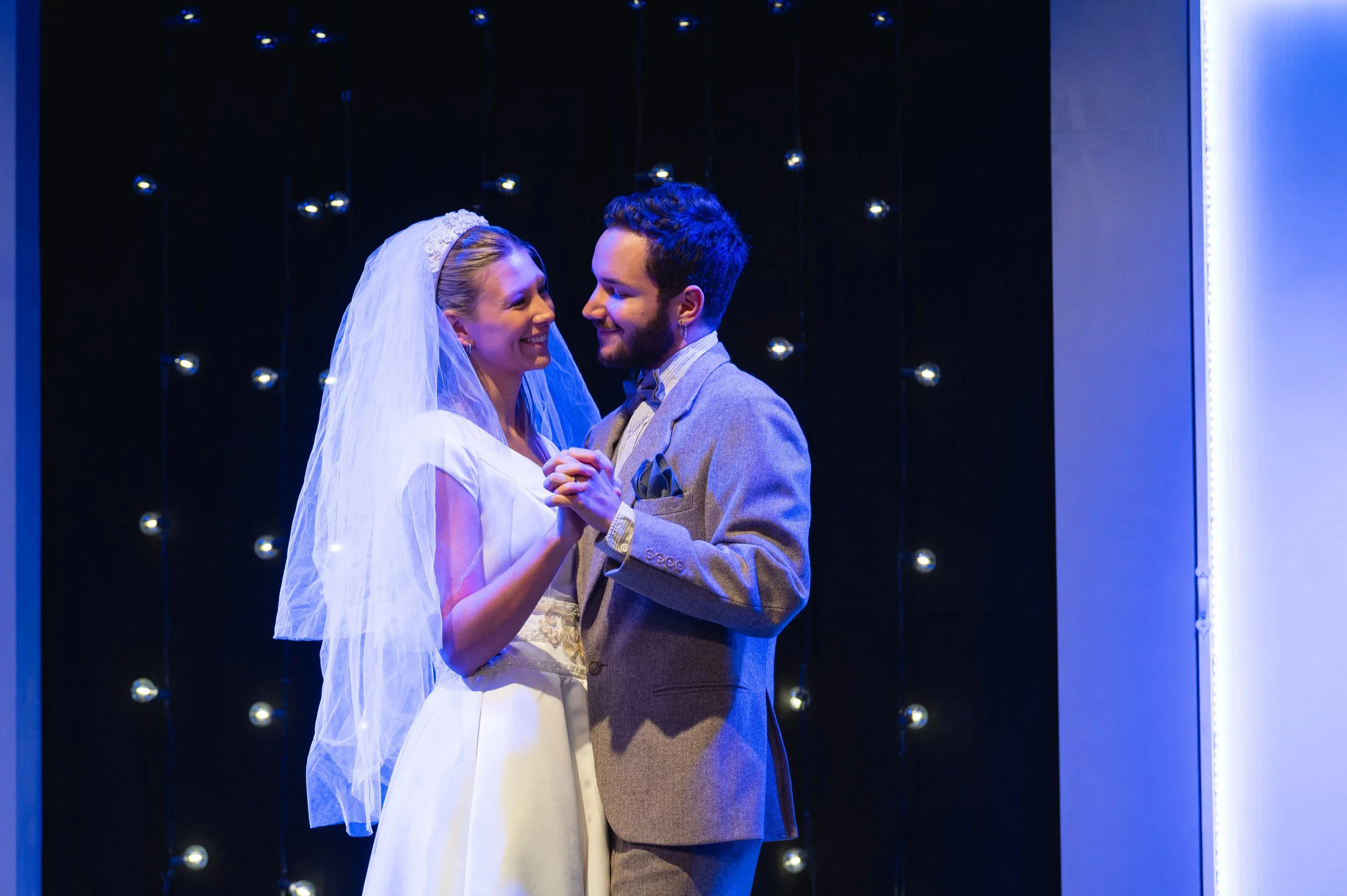 A photograph from the play Significant Other. A bride and groom wedding dance on stage with a dark background and hanging lights, smiling and holding hands.