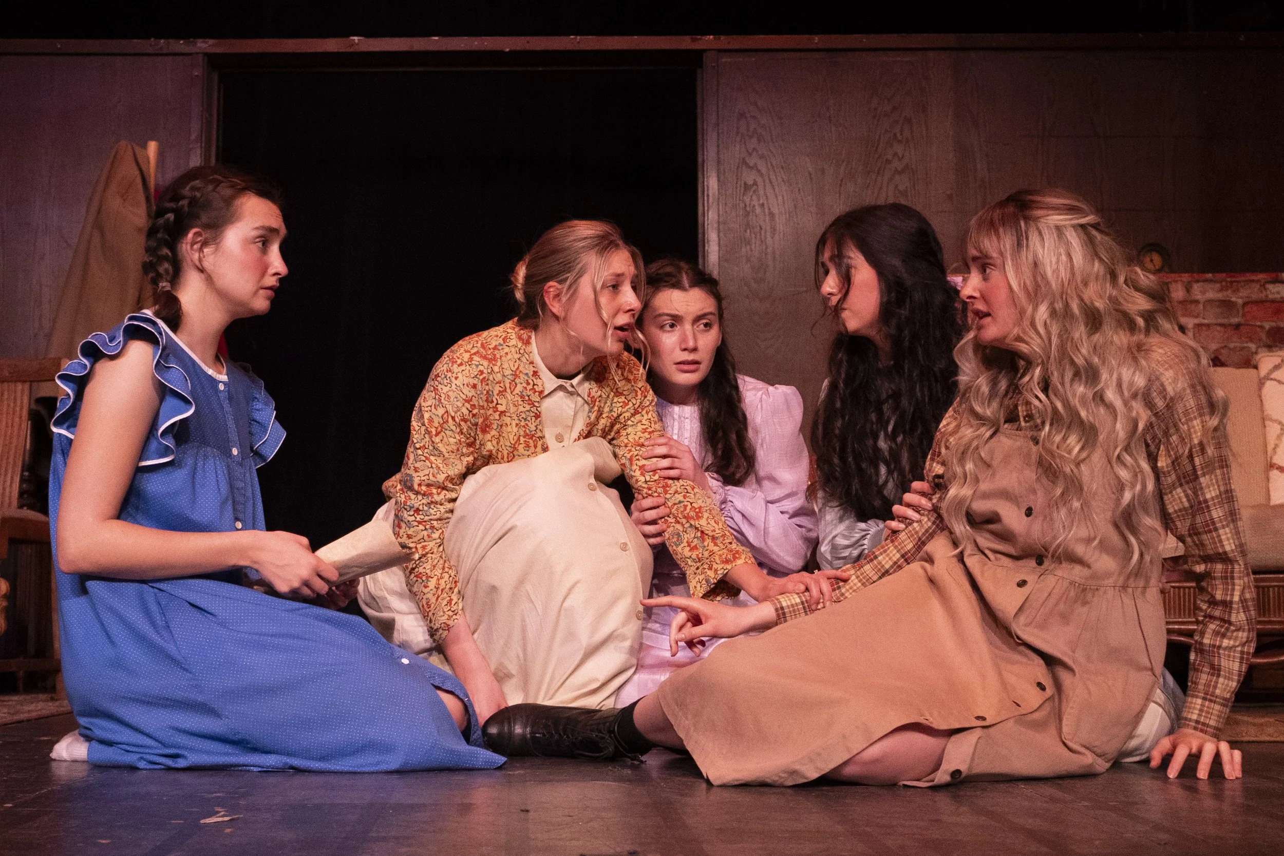 A photograph from the play Little Women. Five women engaging in an emotional discussion on stage, with three women sitting on the floor, and two women kneeling.