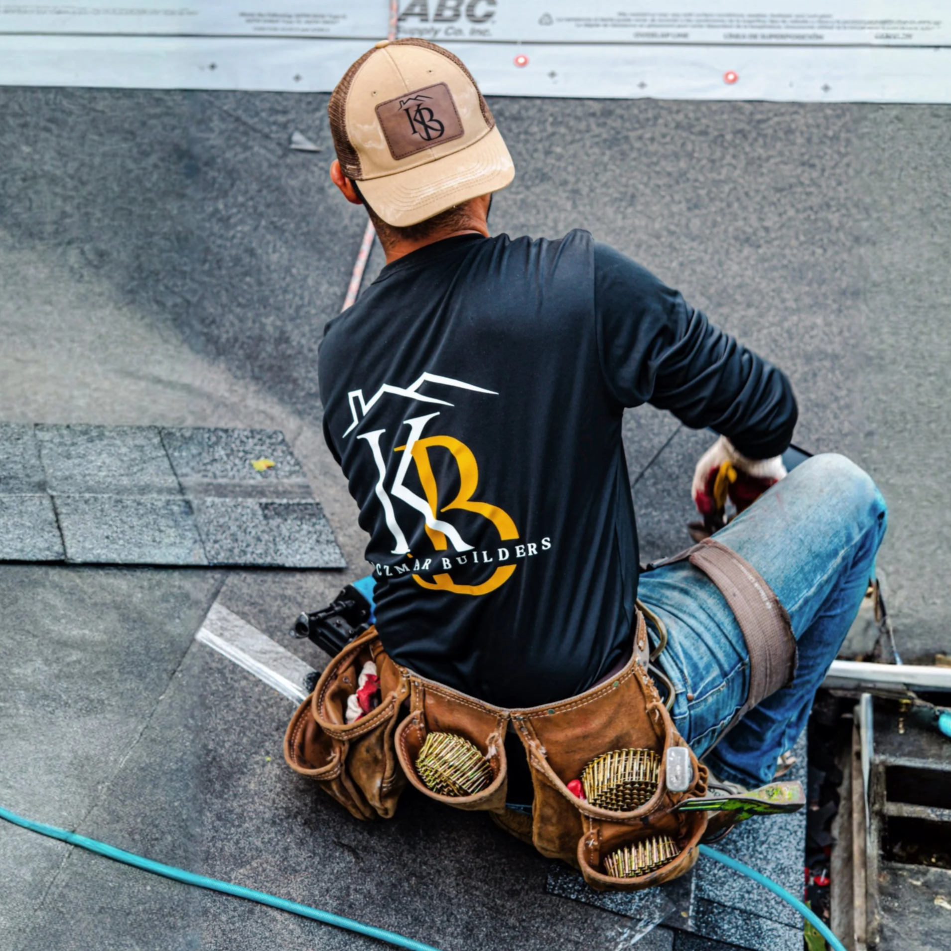 A construction worker installing shingle roofing, wearing a black shirt with a logo, a brown tool belt with various tools, and a beige cap.