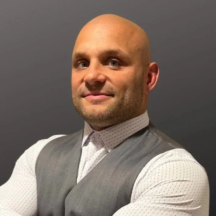 Bald man in a white dotted shirt and gray vest crossing his arms, standing against a gradient gray background.
