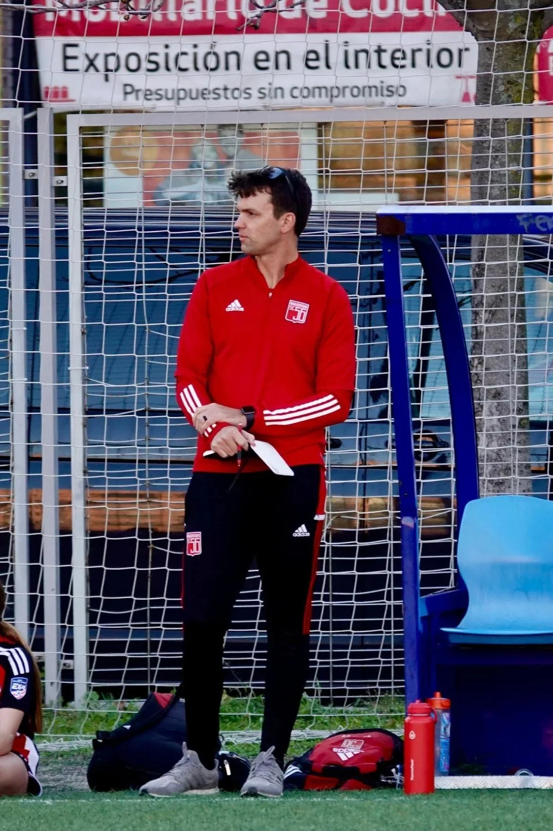 A man standing on the sidelines of a soccer field, dressed in athletic wear with a red Adidas jacket and black track pants, holding a paper, with a backpack and water bottles nearby, behind a fence with a sign in Spanish.