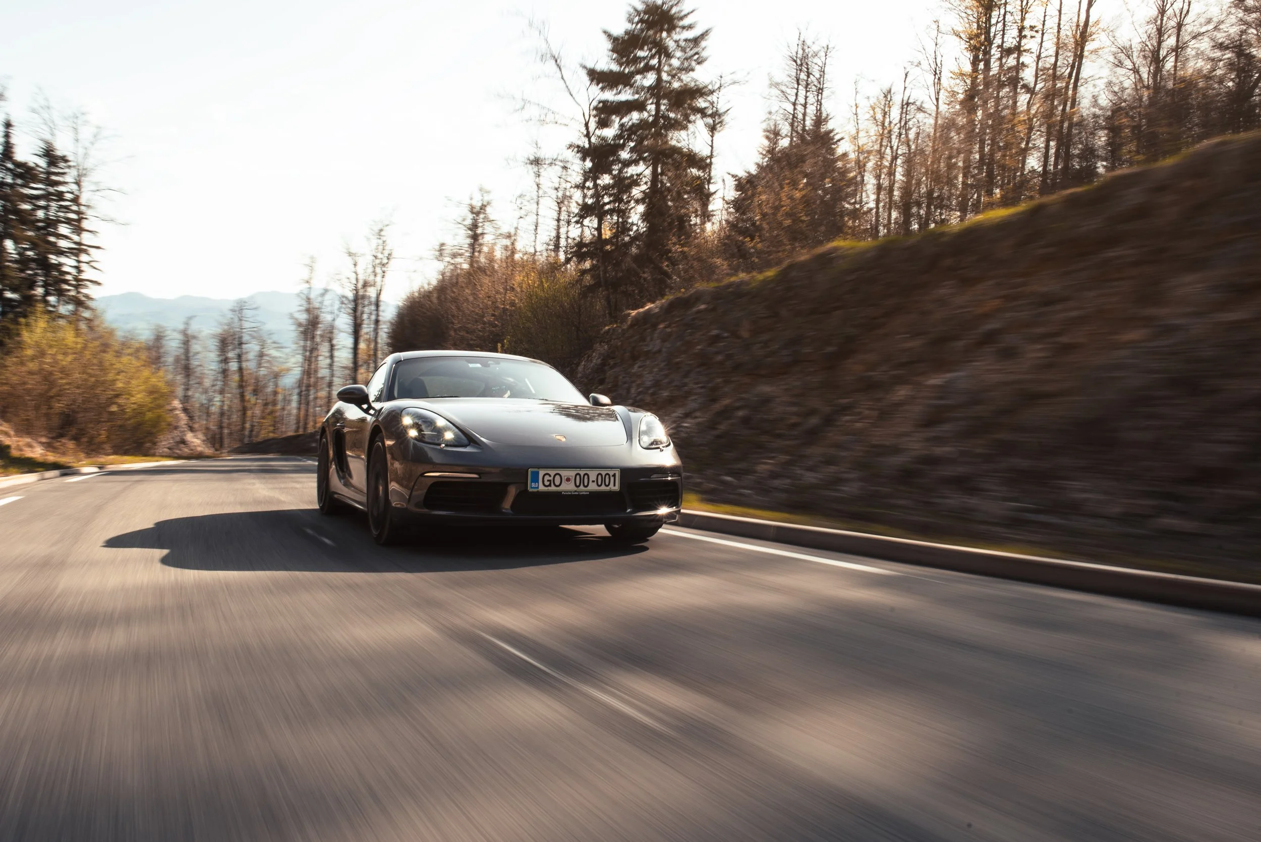 A black sports car driving on a winding mountain road with trees and hills in the background.