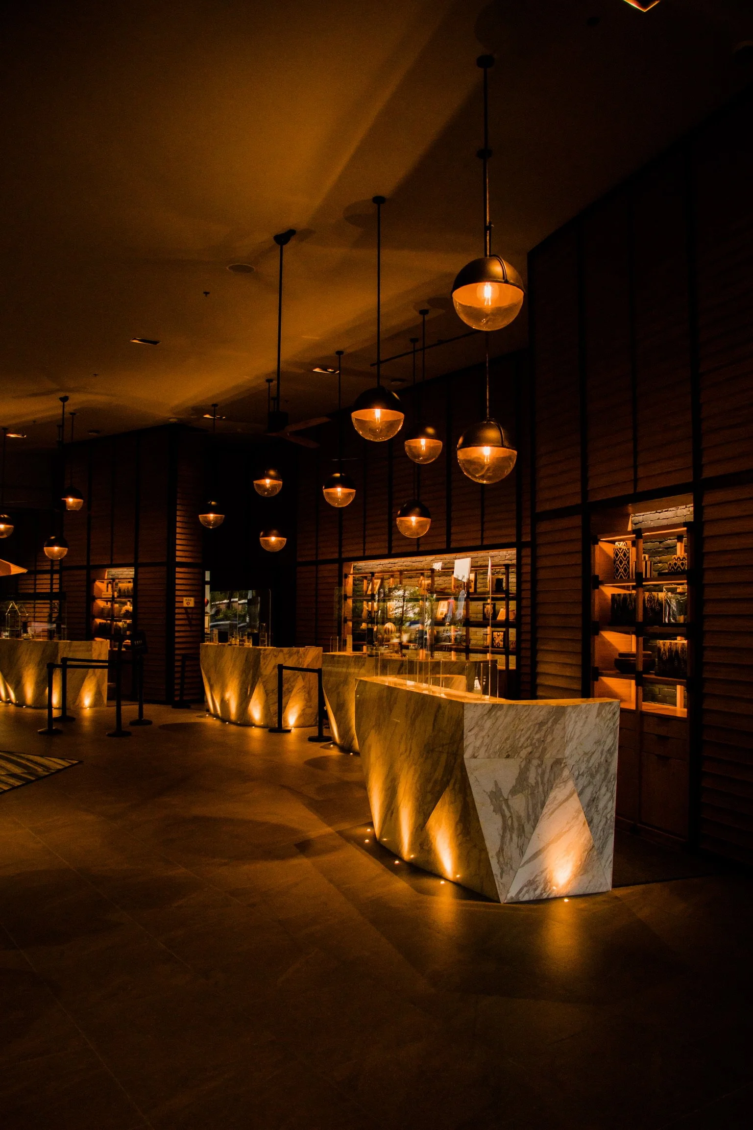 A dimly lit hotel front desk area with illuminated marble counters and modern pendant lights hanging from the ceiling.