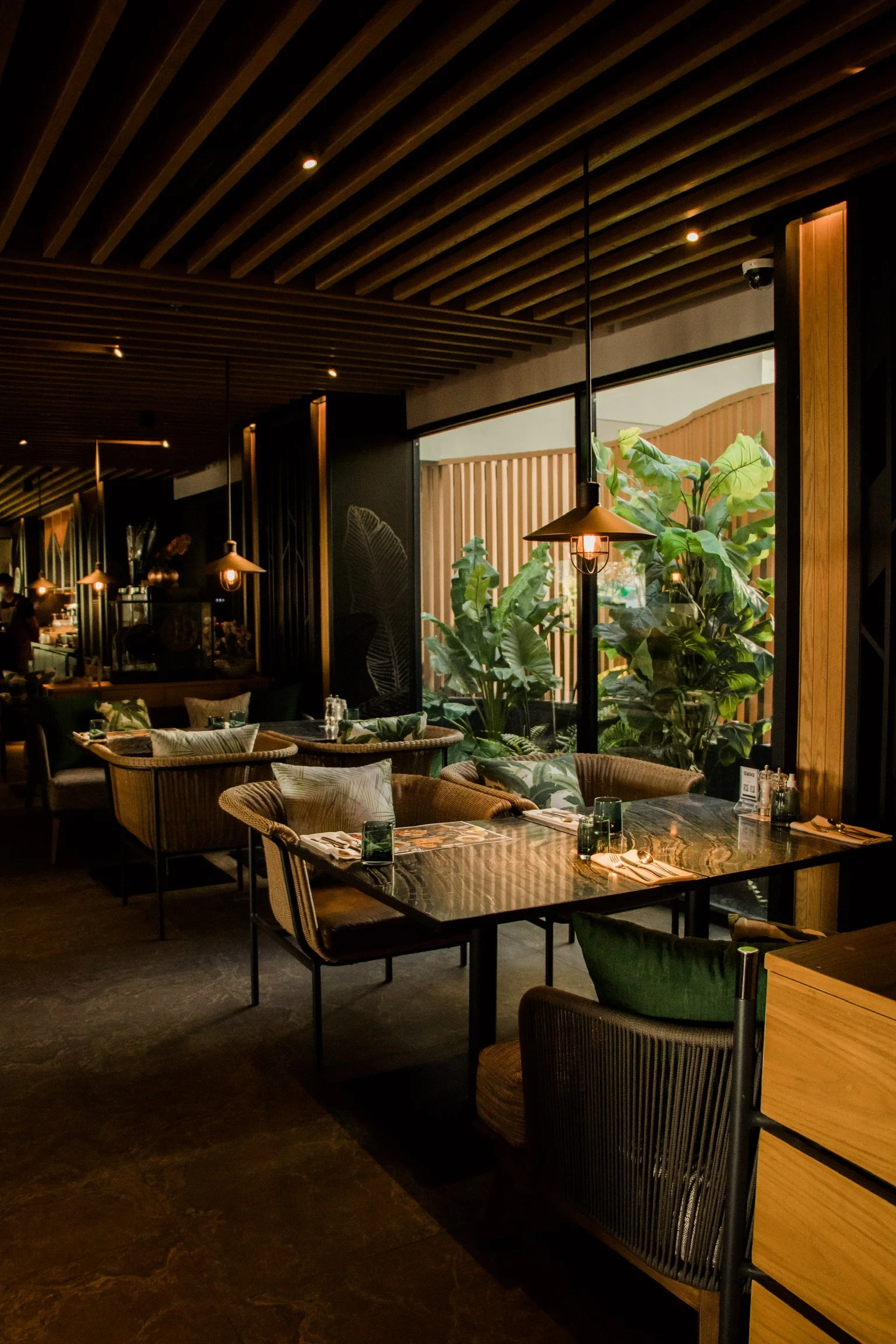 Interior of a restaurant with wooden ceiling beams, illuminated pendant lights, a large window with green plants outside, and tables set with napkins, glasses, and cutlery.