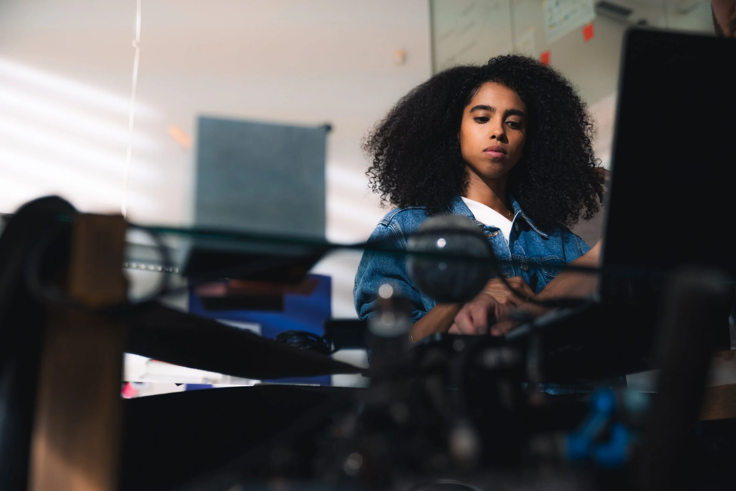 Young woman with curly black hair working on a laptop in an office or classroom setting.