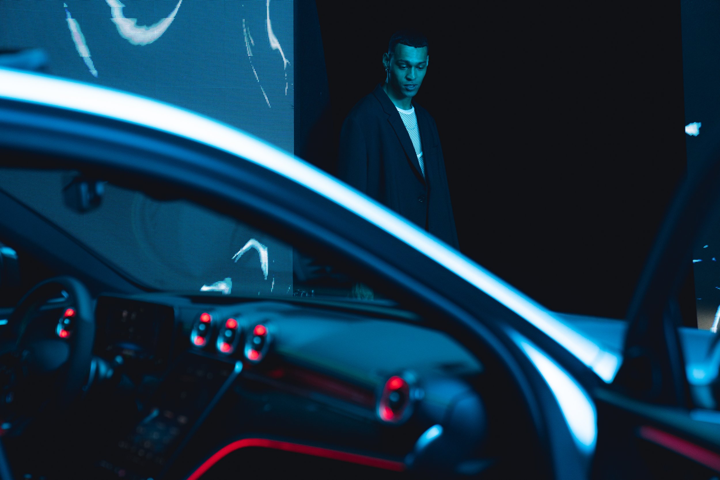 A man standing in a dark room with a blue LED light illuminating his face, viewed from inside a car.