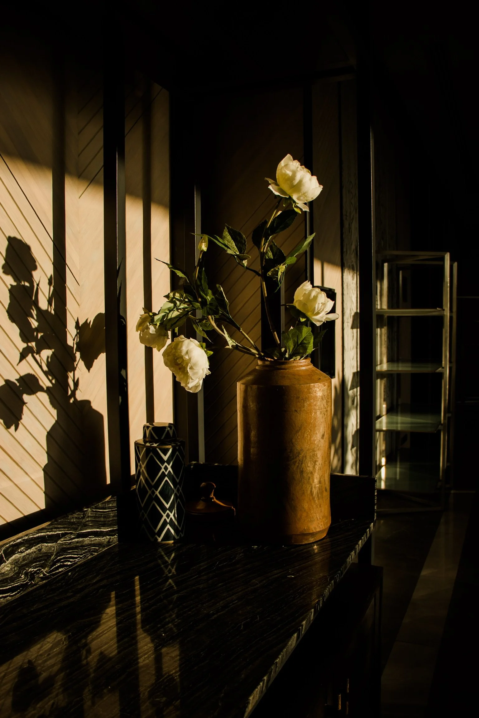 A flower arrangement with white flowers in a large brown vase on a dark marble table, with shadows cast on a wooden wall.