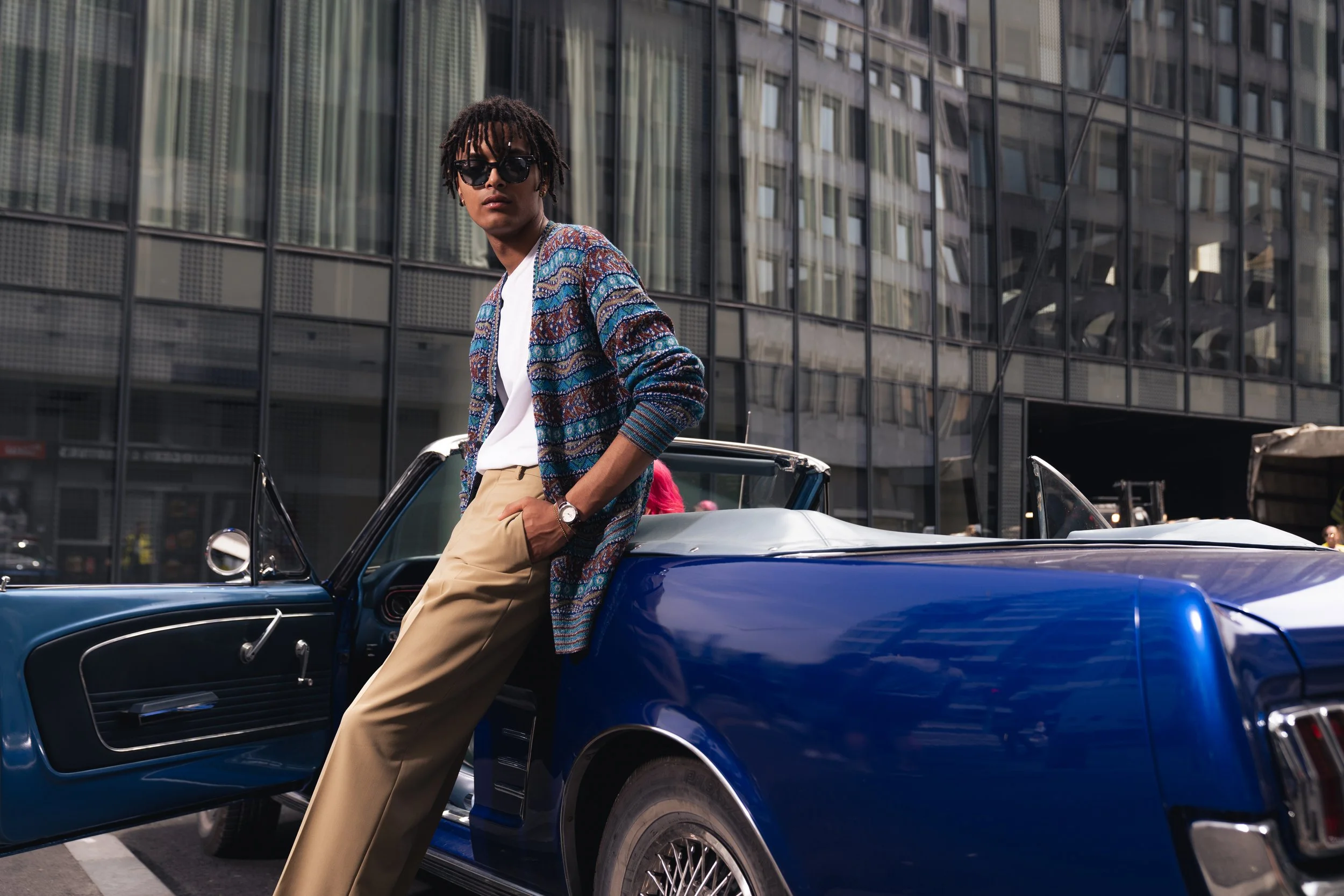 A young man with sunglasses dressed in a patterned blazer, white shirt, and khaki pants leaning against a vintage blue convertible car in an urban setting.