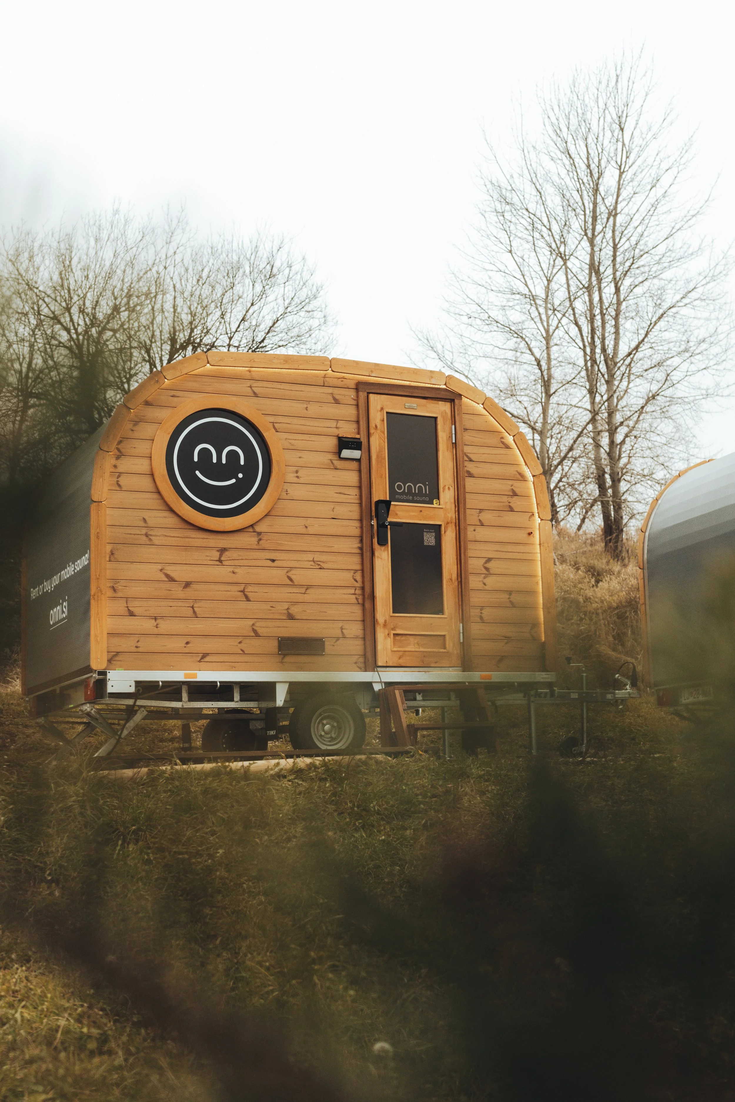 A small wooden mobile sound studio trailer with a smiling circular logo on the front, surrounded by leafless trees.