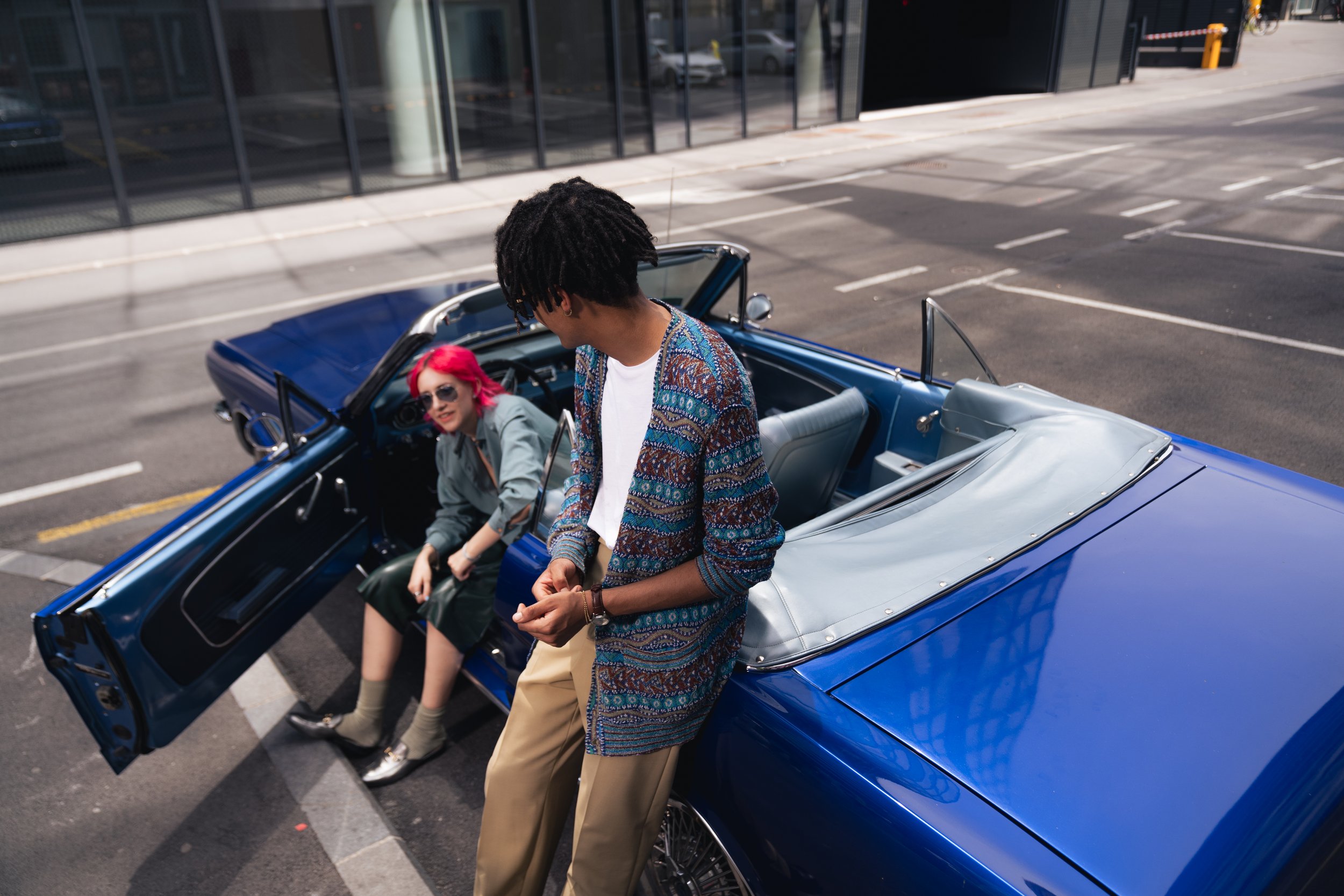 A man in a colorful patterned sweater leans on a vintage blue convertible car while a woman with bright pink hair sits inside, talking and wearing sunglasses.
