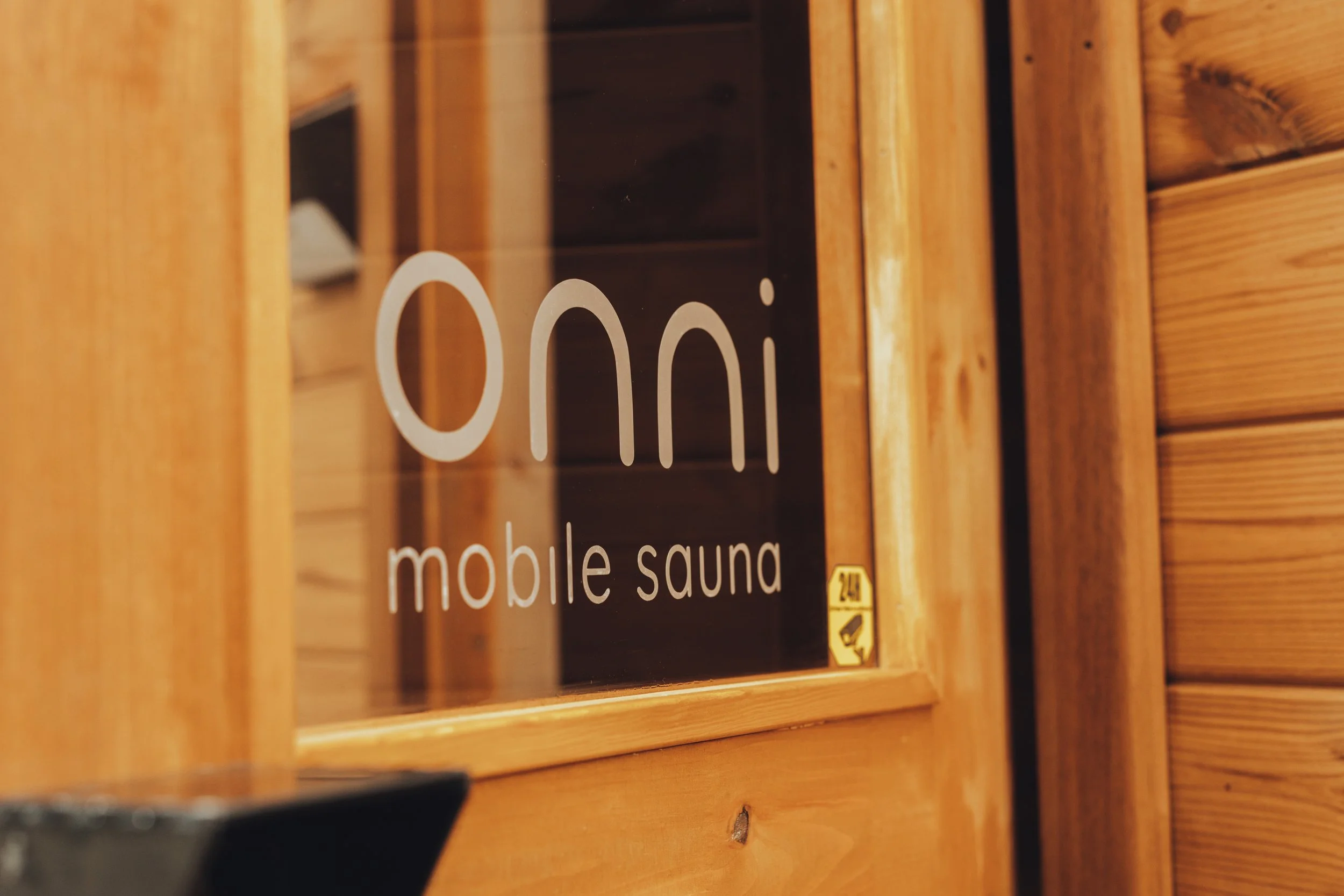 Wooden room with a glass window that has the words 'Oni mobile sauna' written on it and a small yellow sticker in the bottom right corner.