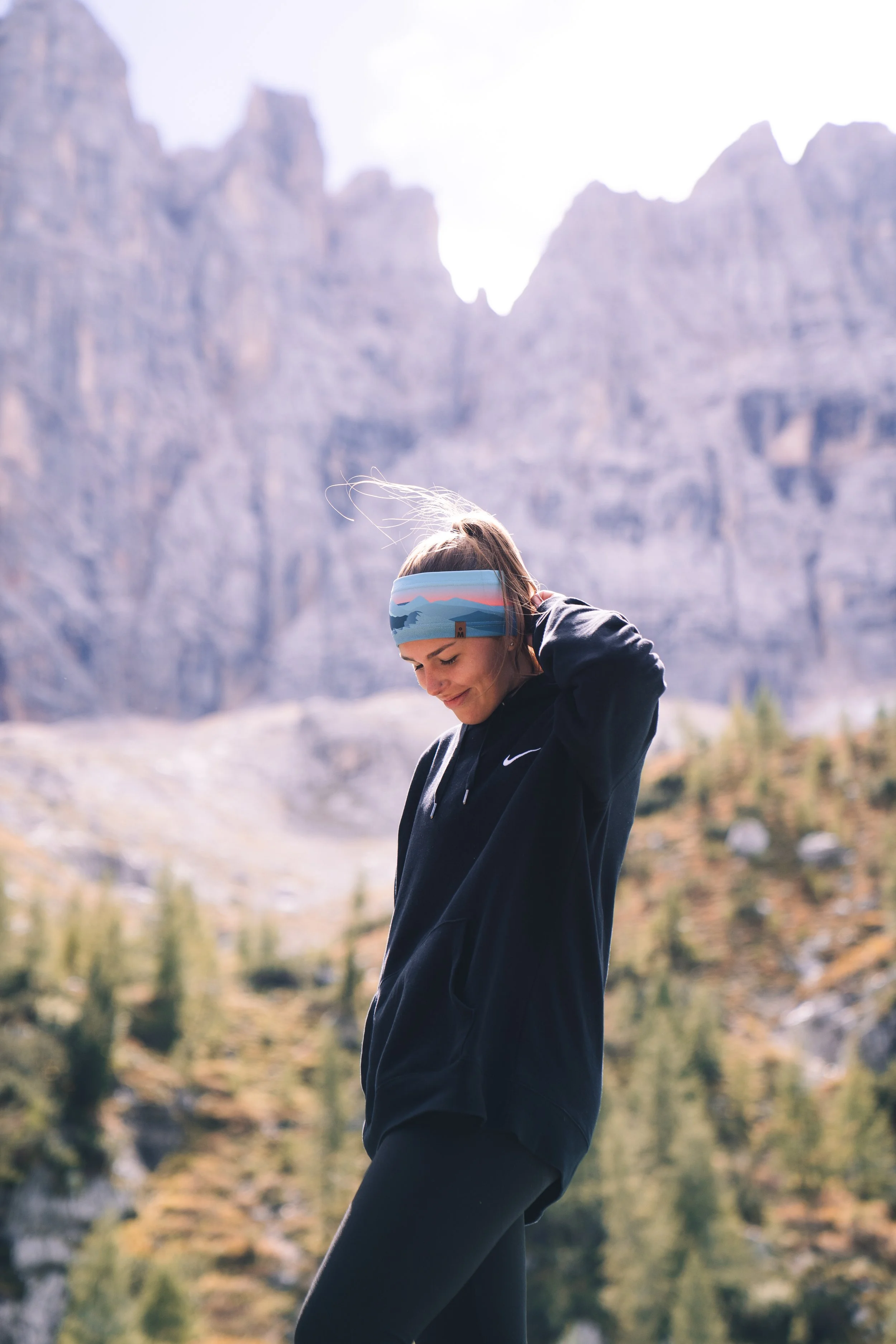 A woman in athletic clothing adjusting her hair outdoors with mountains in the background.