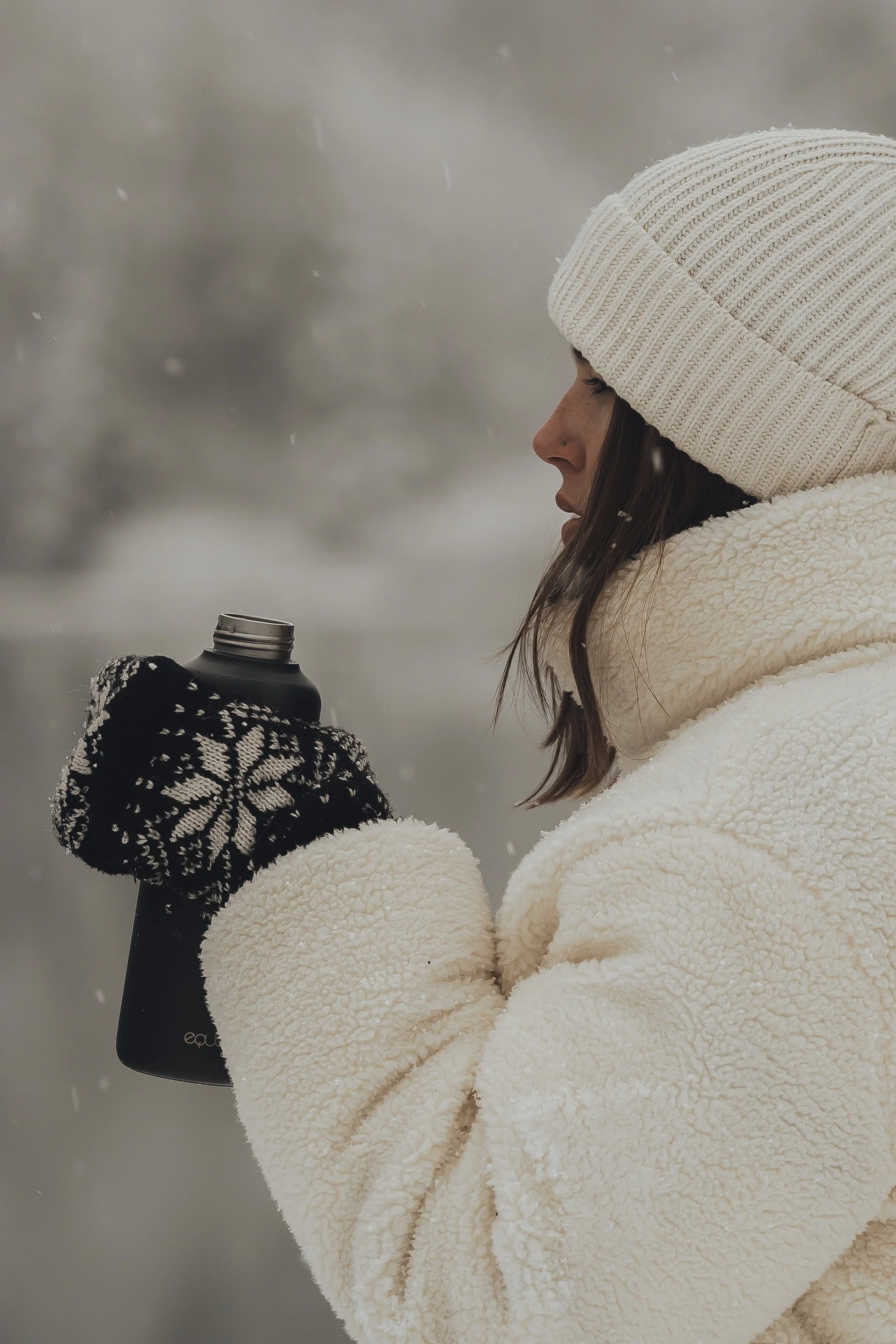 A woman dressed in winter clothing, including a cream-colored fleece jacket and a knit beanie, holding a black thermal water bottle outdoors in snowy weather.