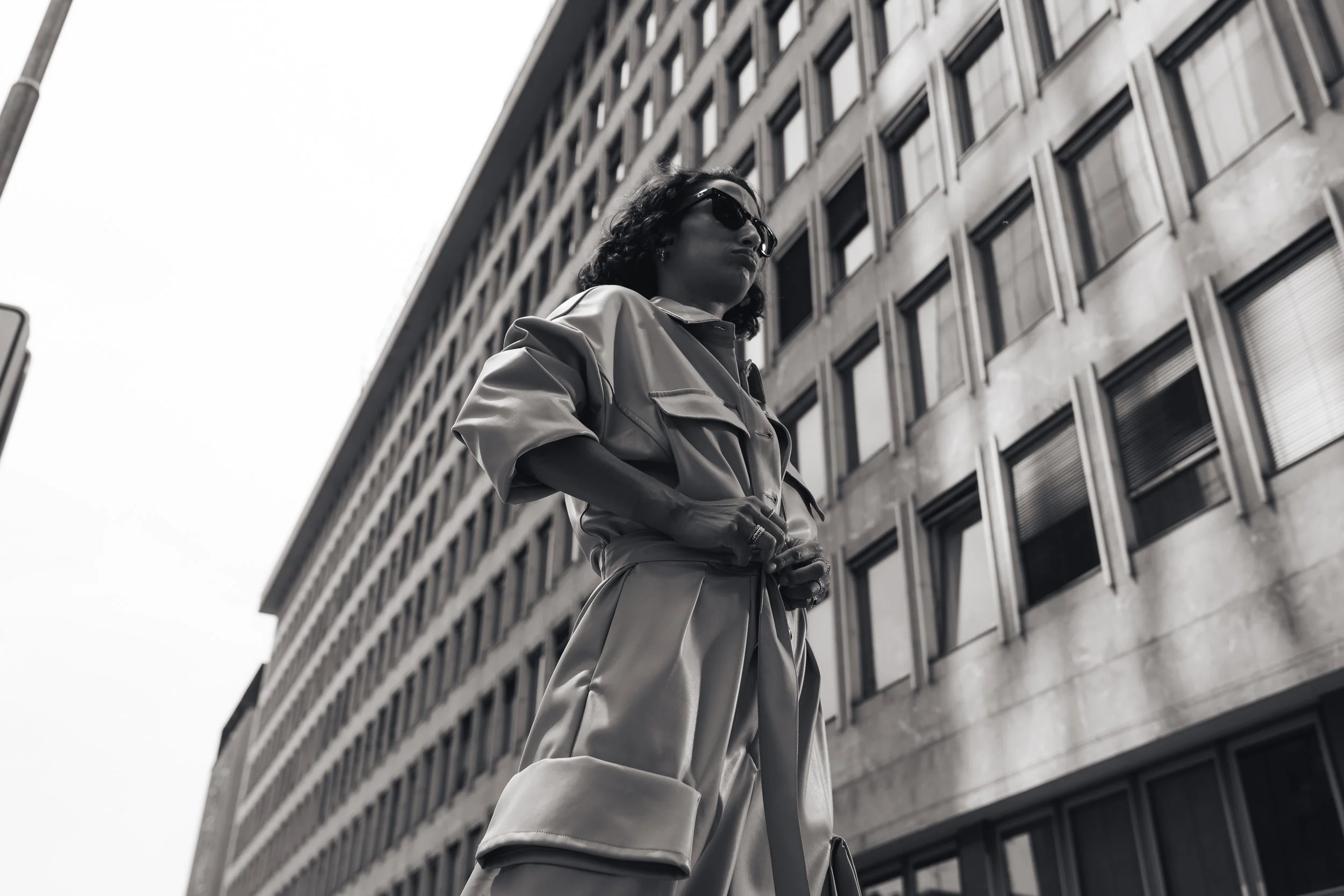 A woman wearing sunglasses and a loose-fitting jumpsuit stands in front of a modern office building, looking to her left.