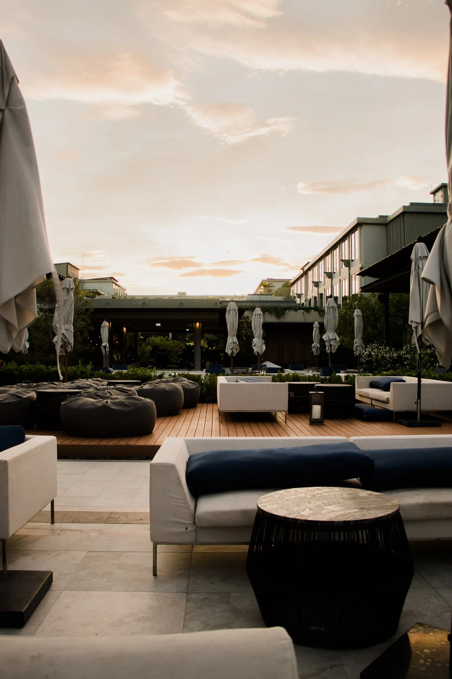 Outdoor lounge area with white, black, and navy seating, bean bags, umbrellas, and a wooden deck, under a partly cloudy sky during sunset.