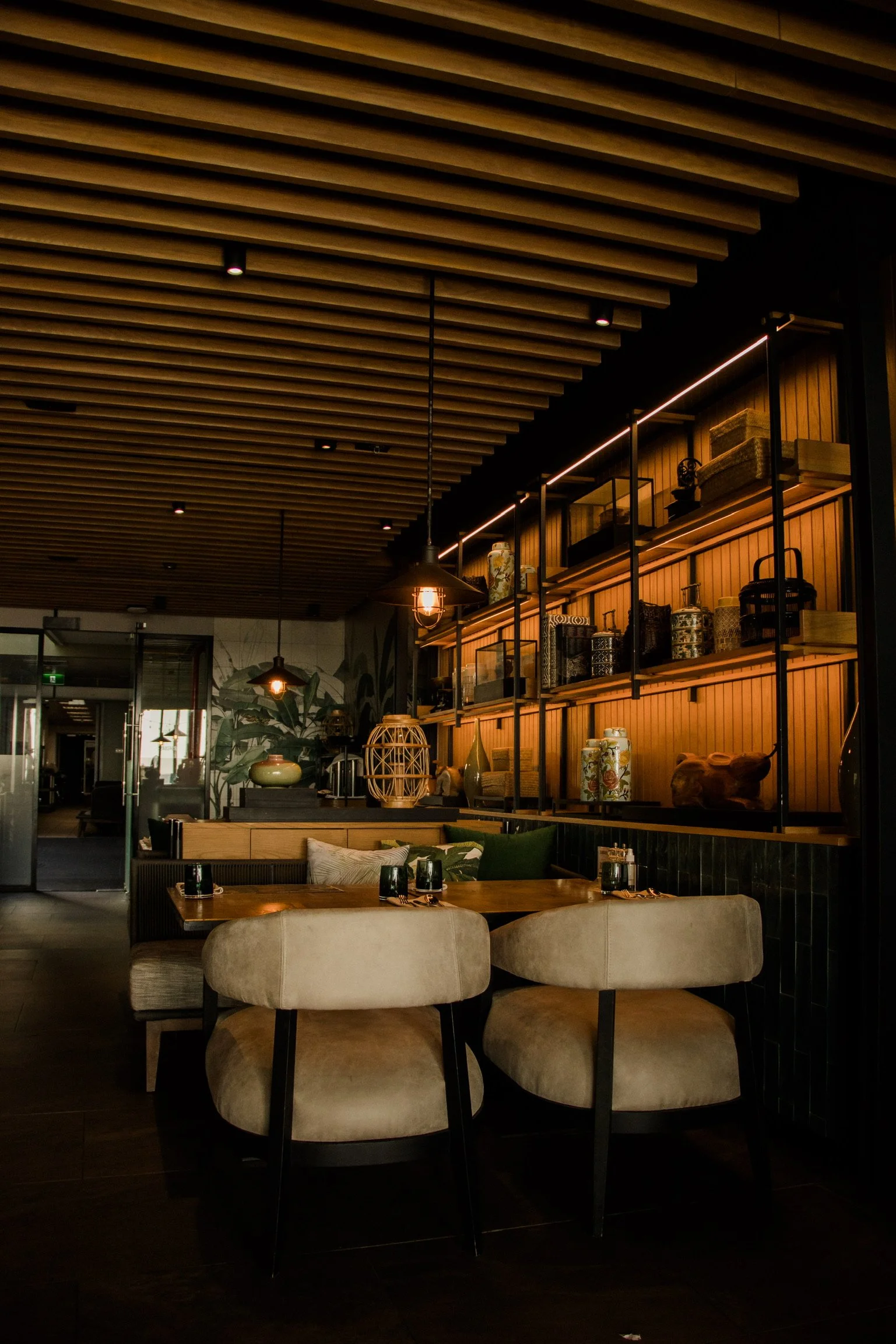 Cozy restaurant interior with wooden ceiling, decorative shelves with vases and boxes, booth seating, and ambient lighting.