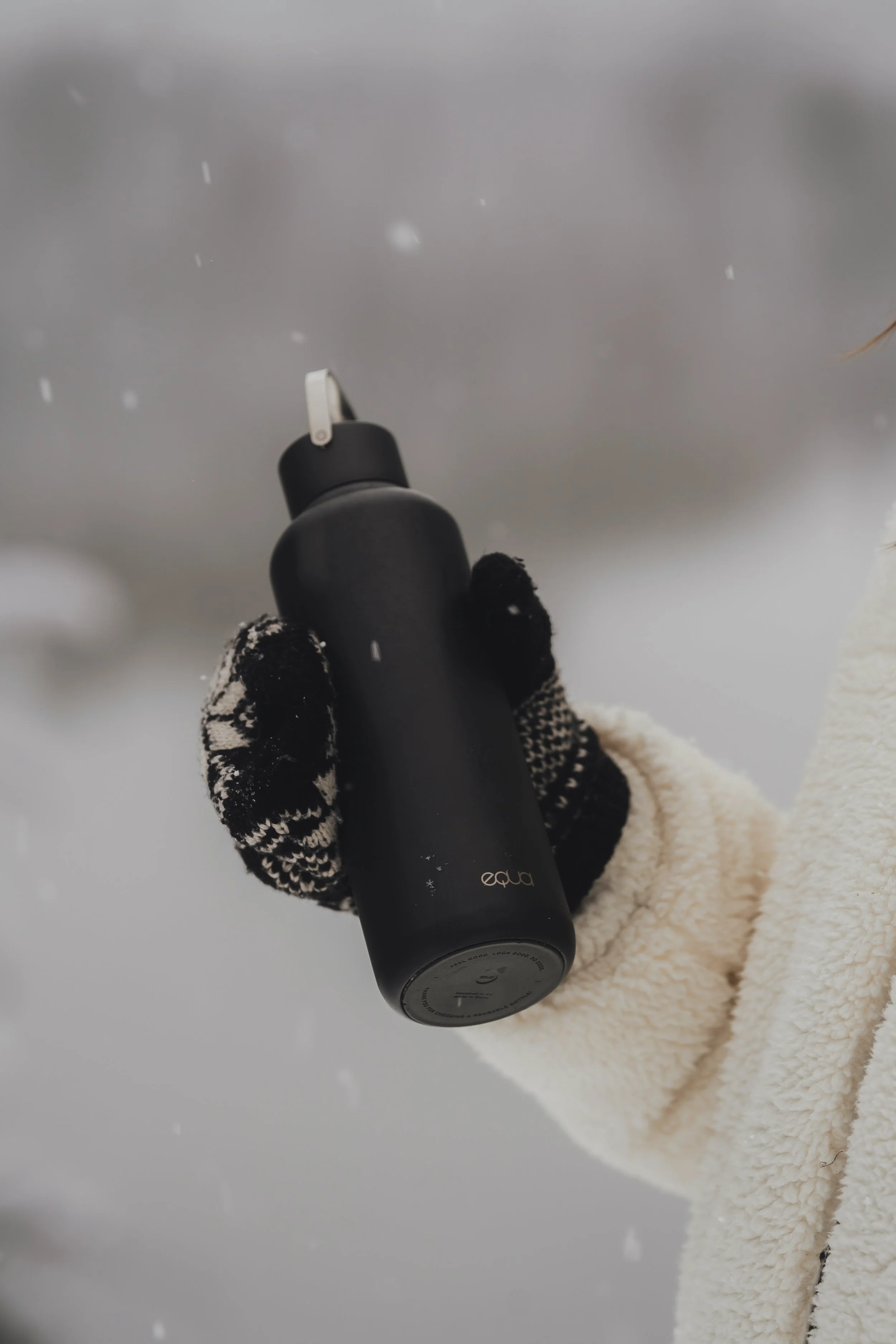 Person wearing black and white patterned gloves holding a black Eucalyptus natural deodorant bottle outdoors in a snowy environment.