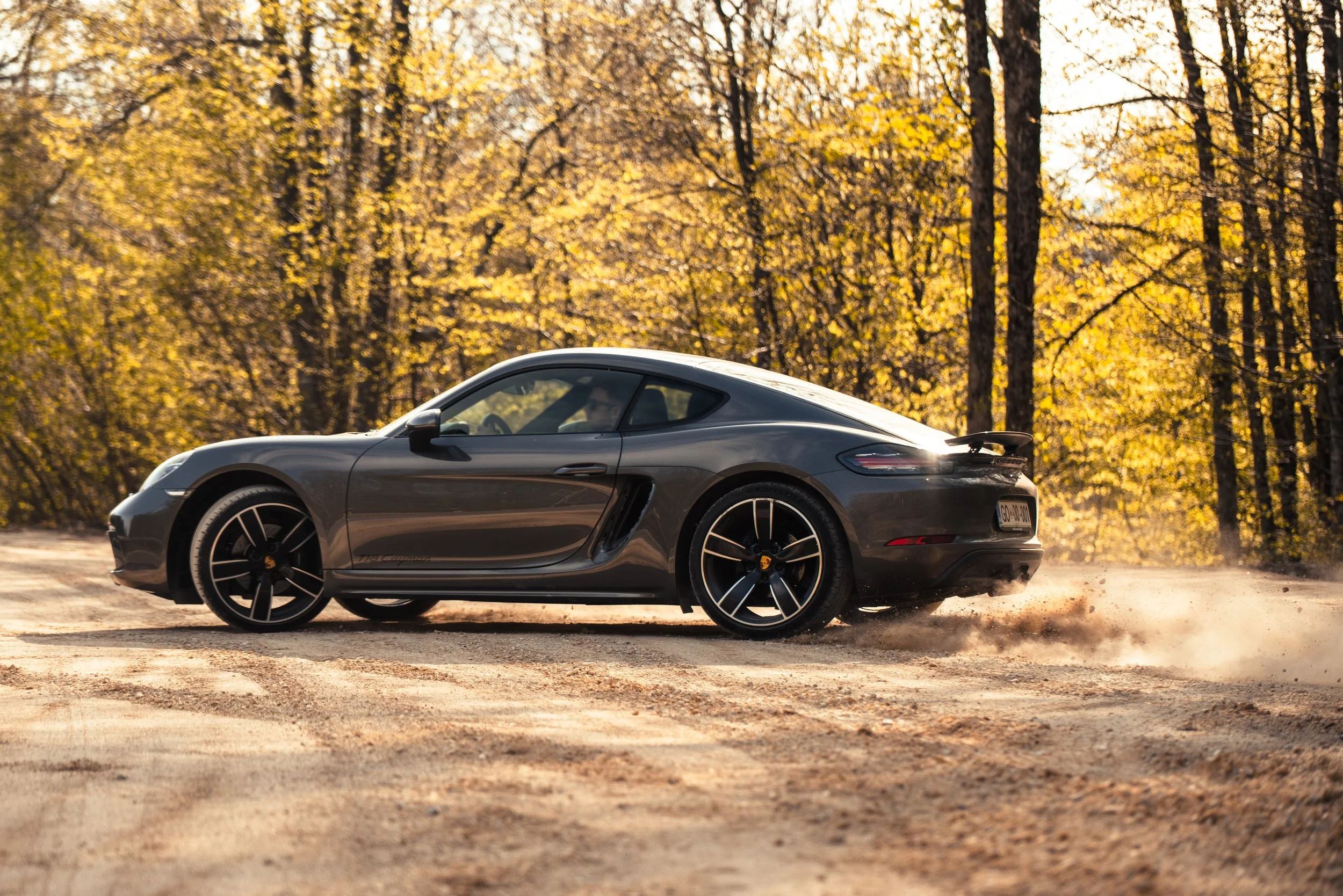 A gray sports car driving on a dirt road in a wooded area during autumn, kicking up dust behind it.