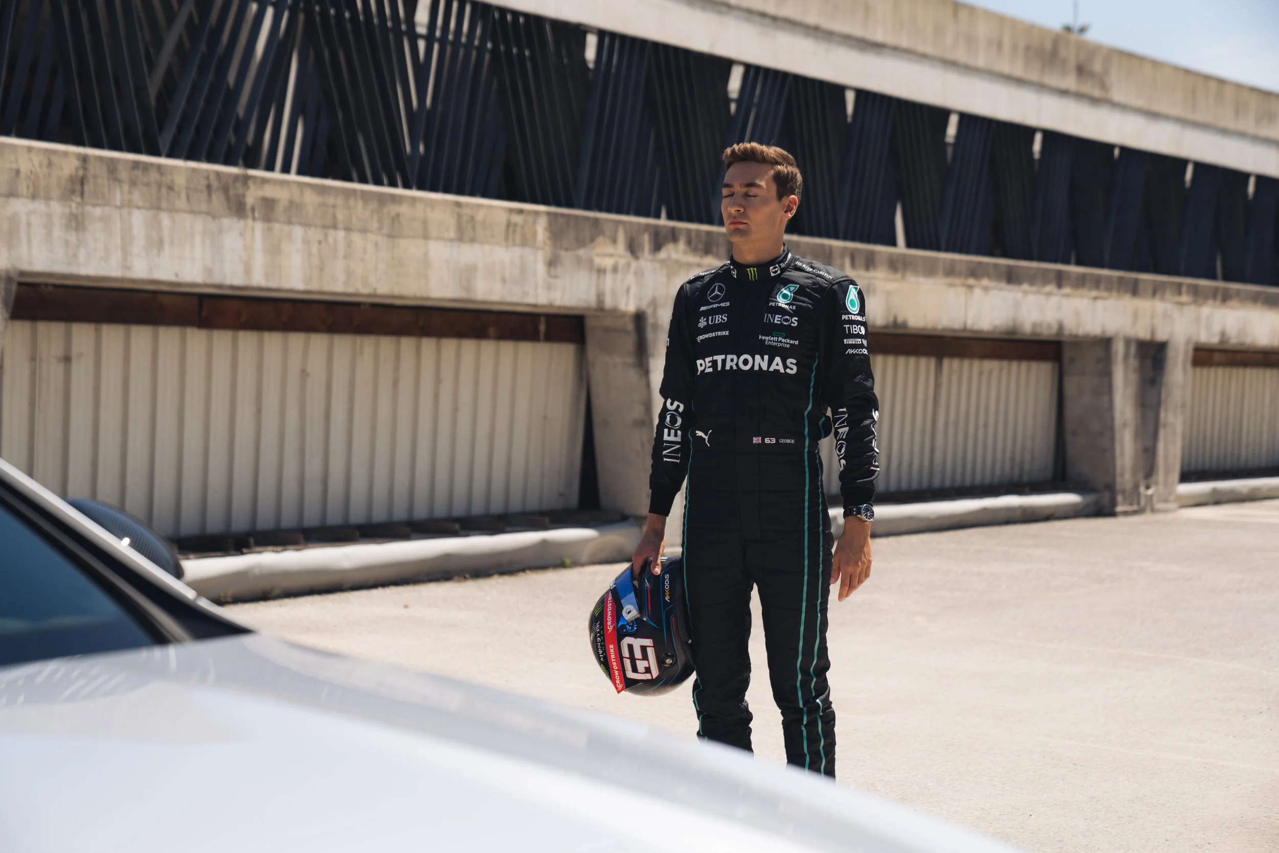 A young male race car driver in a black racing suit standing next to a race track holding a helmet in his right hand, with a concrete structure in the background.