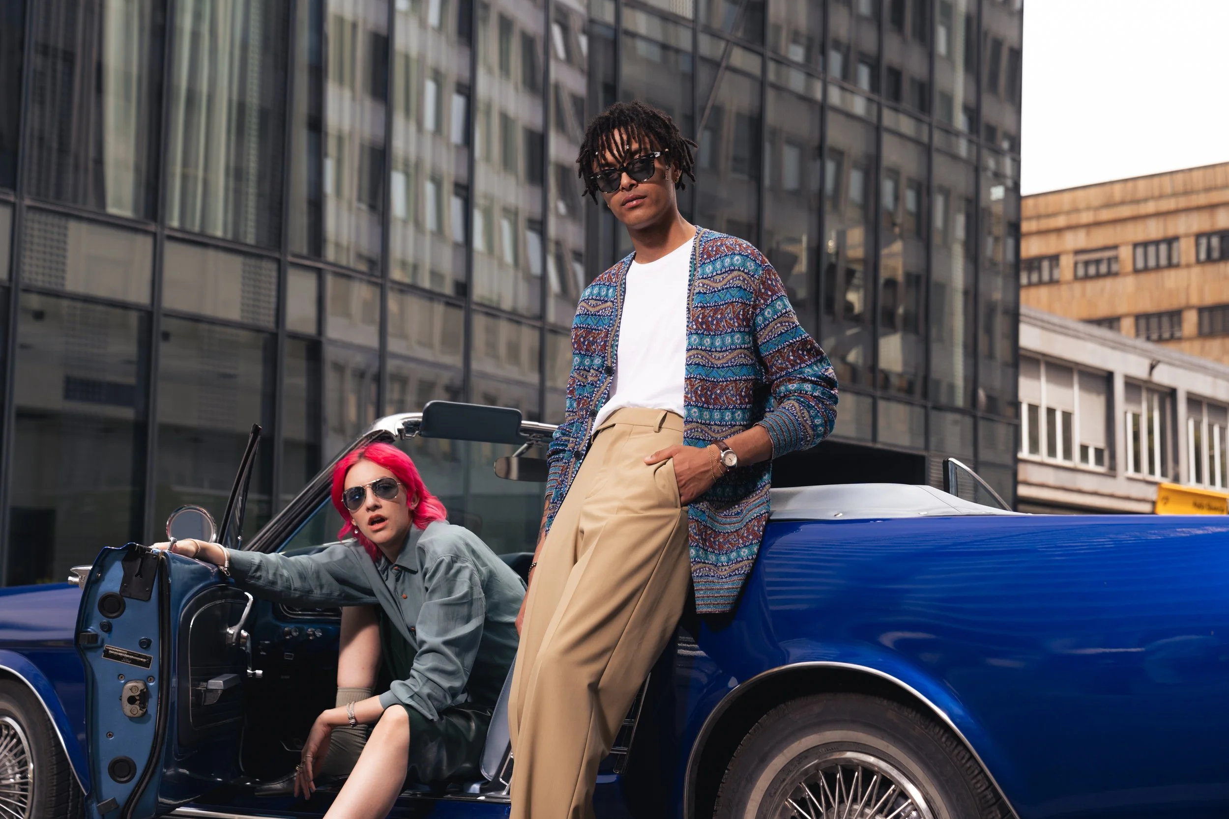 Two fashionable young women with sunglasses and brightly colored hair are posing with a vintage blue convertible car in an urban setting with modern glass buildings.