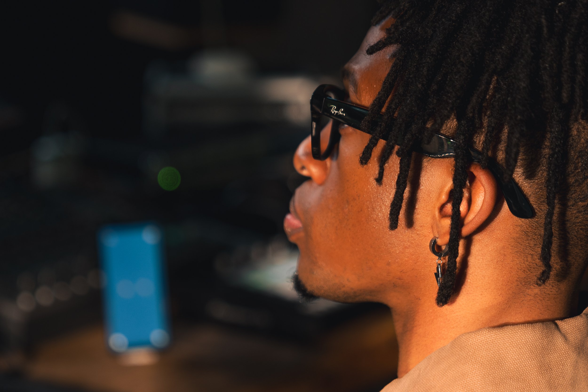 A young man with dreadlocks and glasses looking at a computer screen in a dimly lit environment.