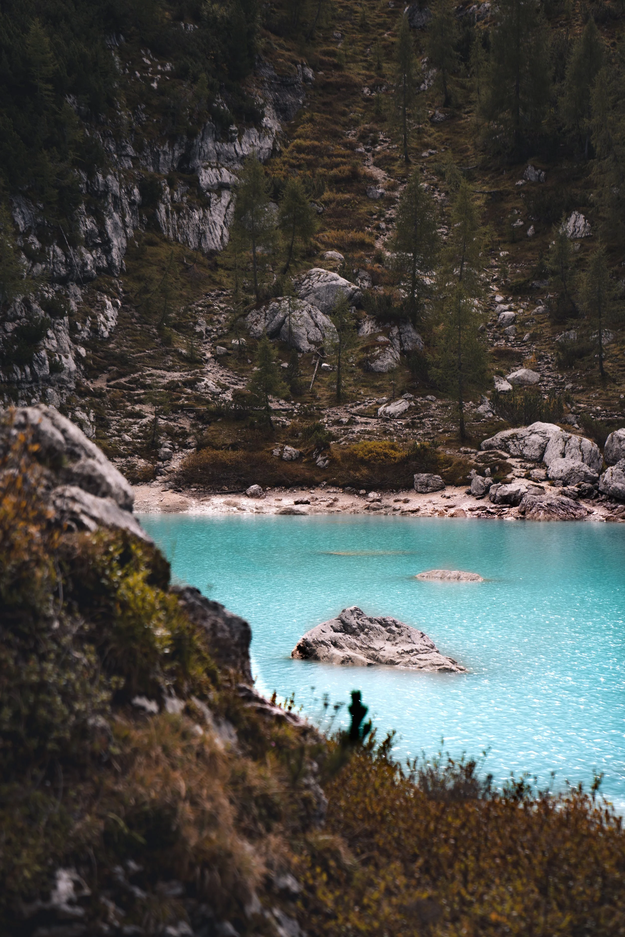 A serene mountain landscape featuring a turquoise lake with rocks, surrounded by dense evergreen trees and rocky cliffs.