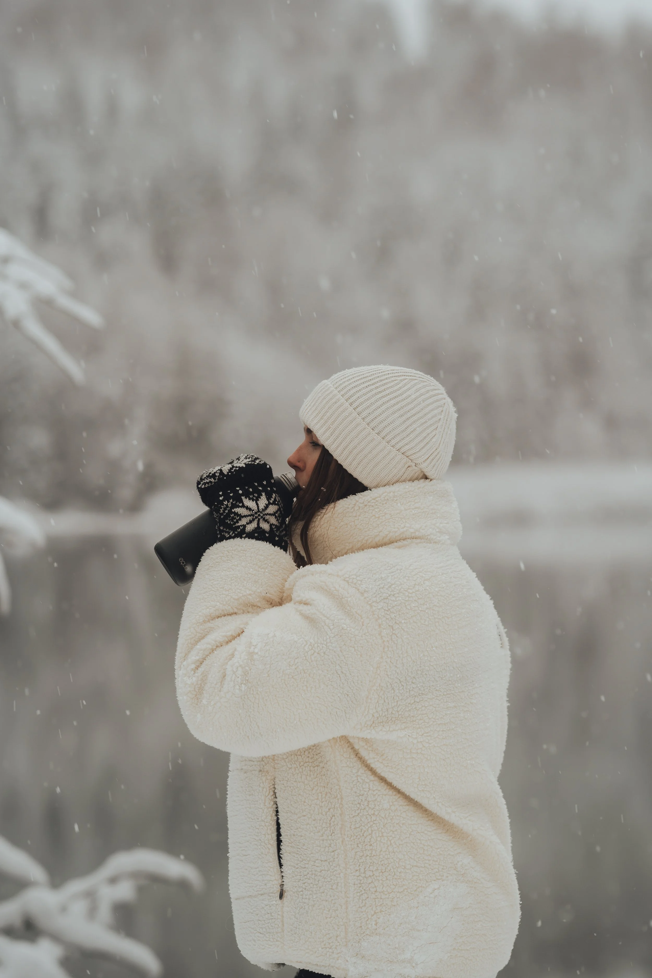 Woman wearing a white knit beanie, cream-colored fleece jacket, and black gloves with snowflake pattern, drinking from a black bottle in a snowy outdoor setting.