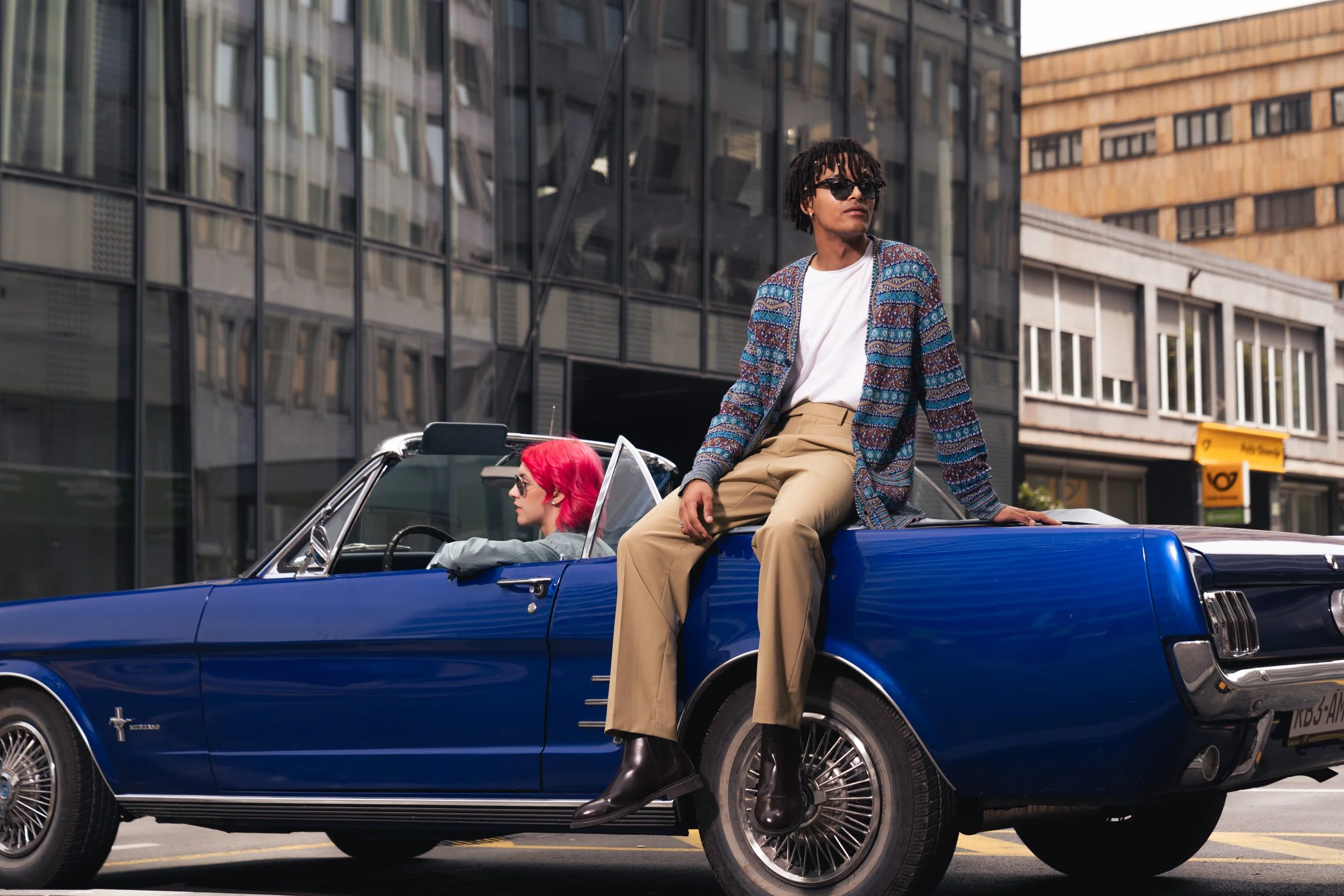 A man with dreadlocks sitting on the trunk of a vintage blue convertible car with a woman with pink hair driving.