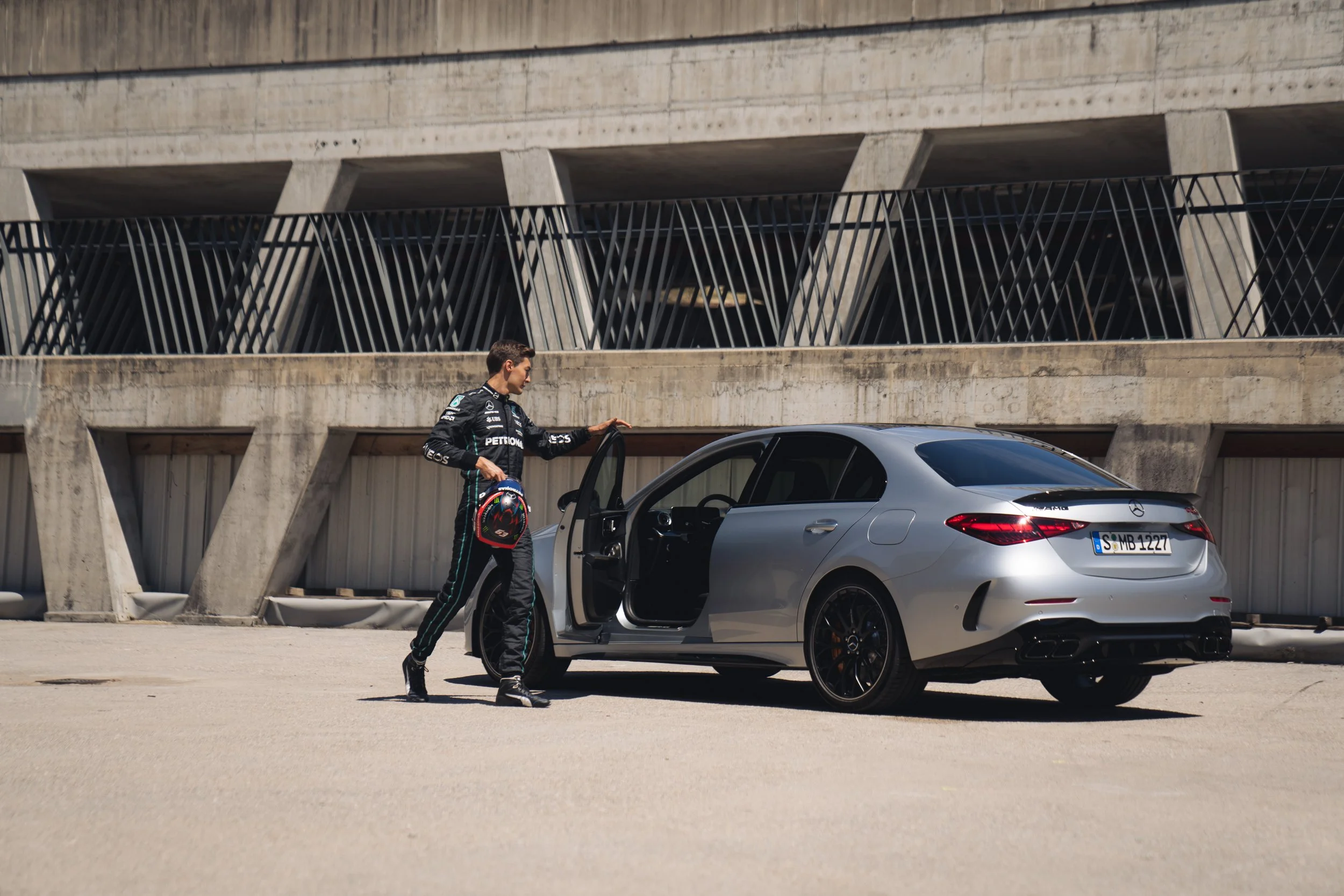 A race car driver in racing suit standing next to a silver Mercedes-Benz sedan in a parking lot.
