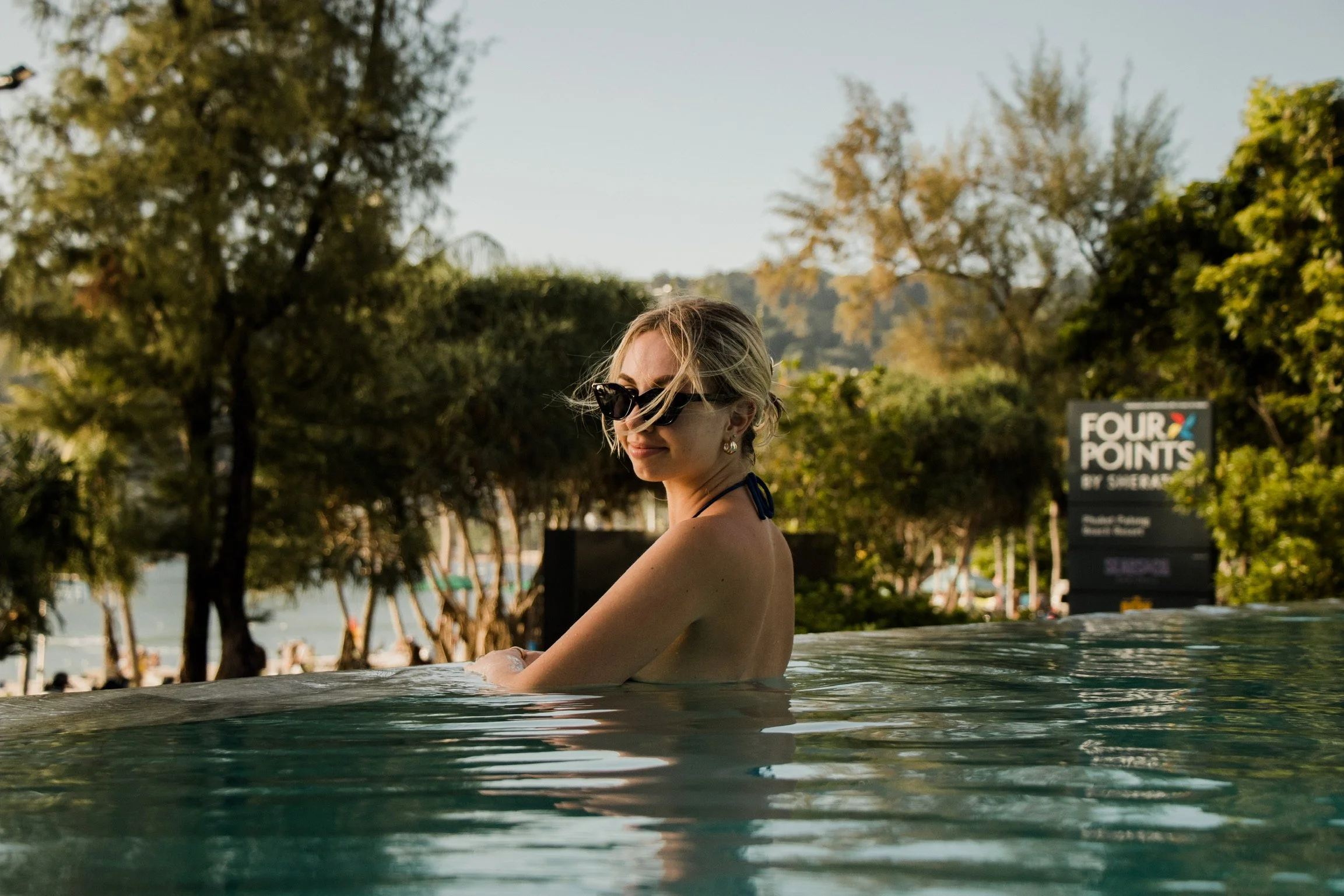 A woman with blonde hair wearing dark sunglasses and earrings, sitting in an outdoor swimming pool with trees and a sign reading "Four Points by Sheraton" in the background.