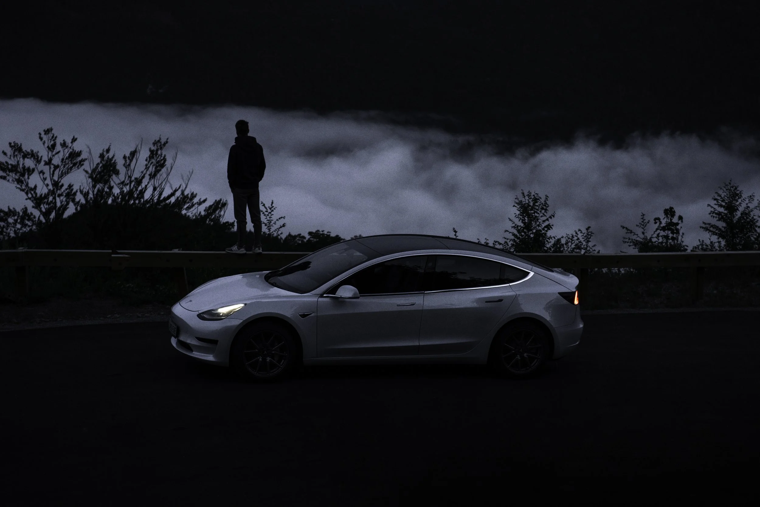 A silver Tesla parked on a dark roadside at night, with a person standing on the guardrail overlooking clouds and fog below.