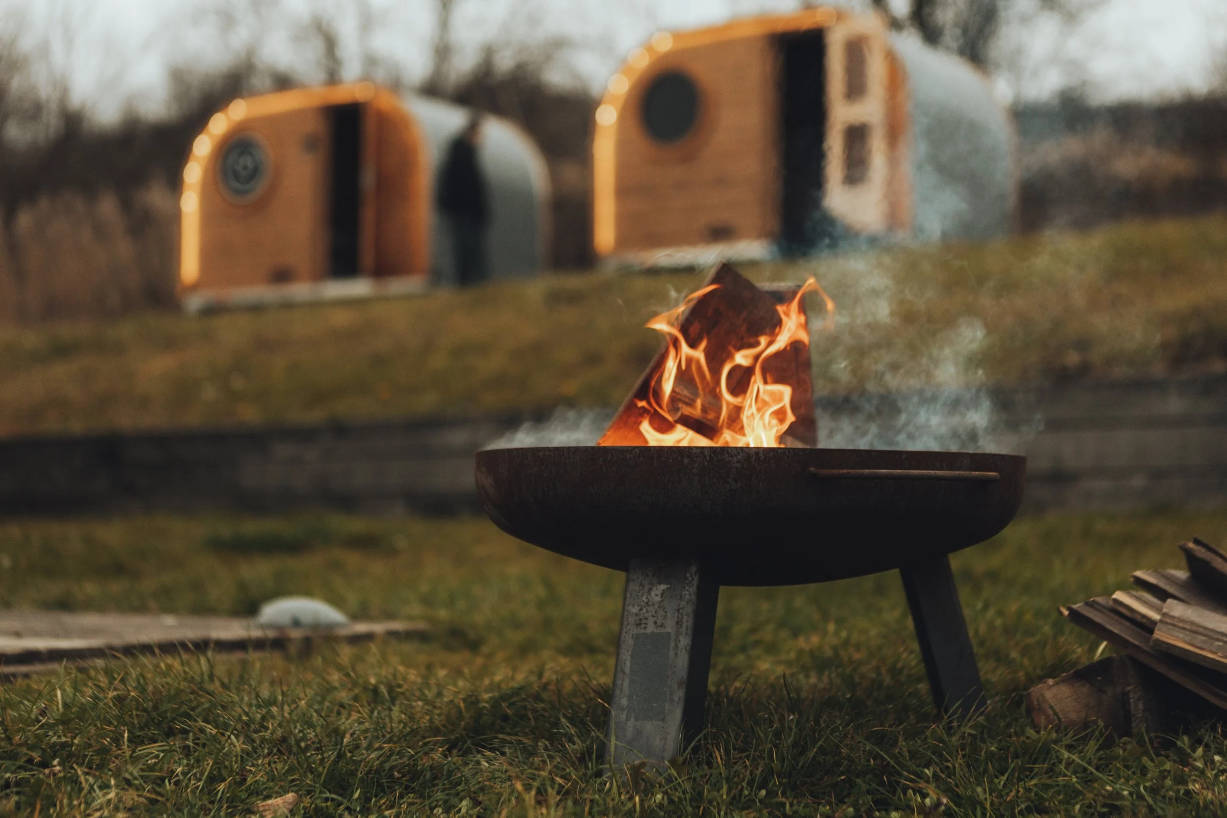 A small fire burning on a portable grill outdoors on grass, with two tiny houses or cabins blurred in the background.