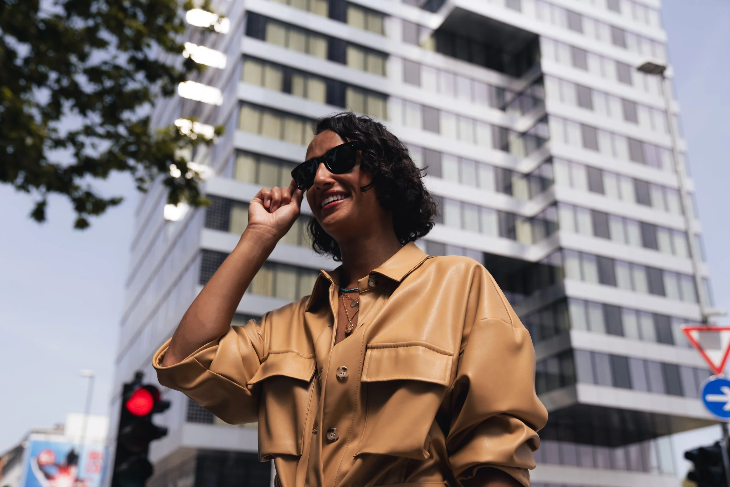 A woman in sunglasses smiling and adjusting her sunglasses in an urban setting with a tall modern building in the background.